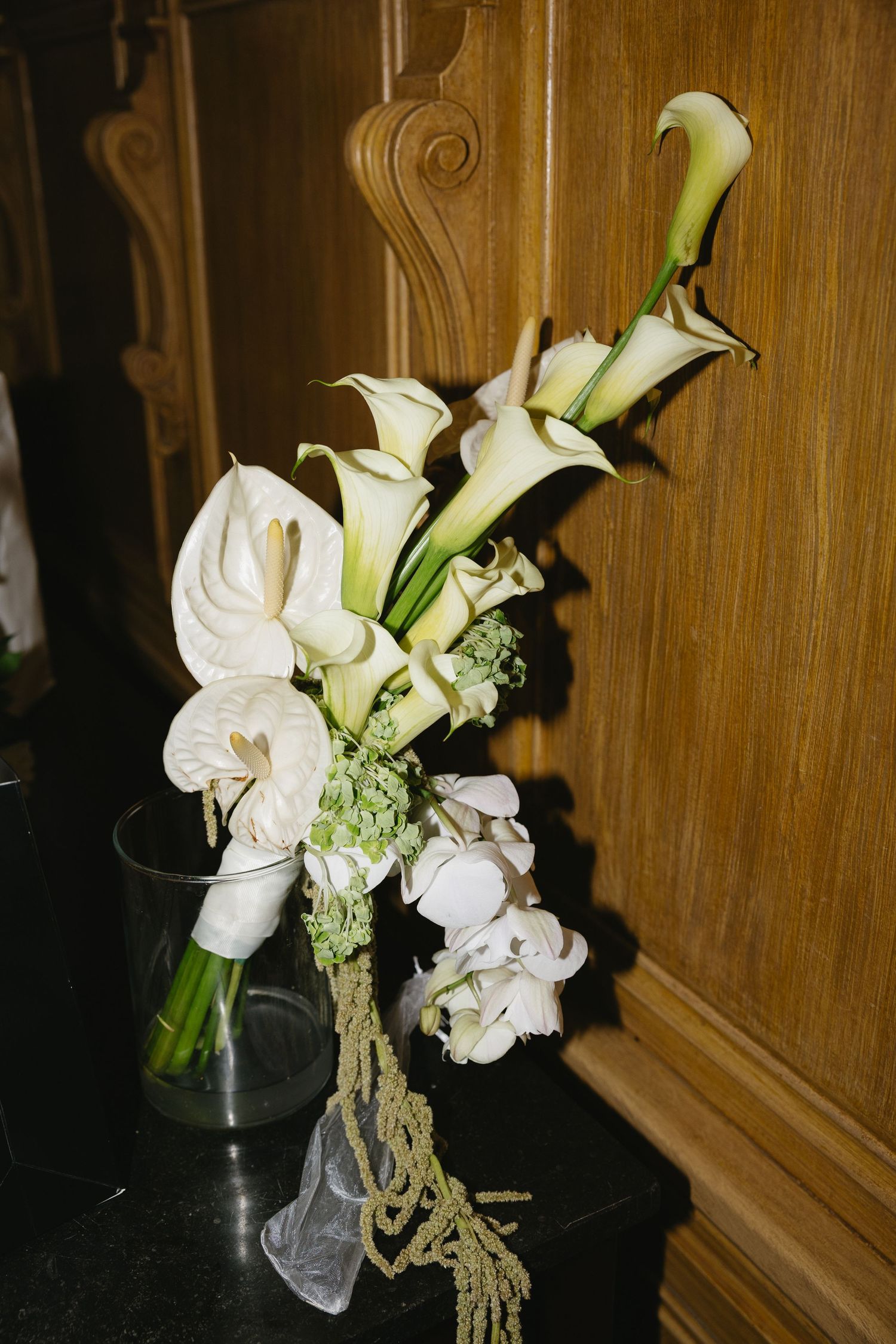 Elegant white calla lily wedding bouquet with green accents displayed in a glass vase on a dark surface.