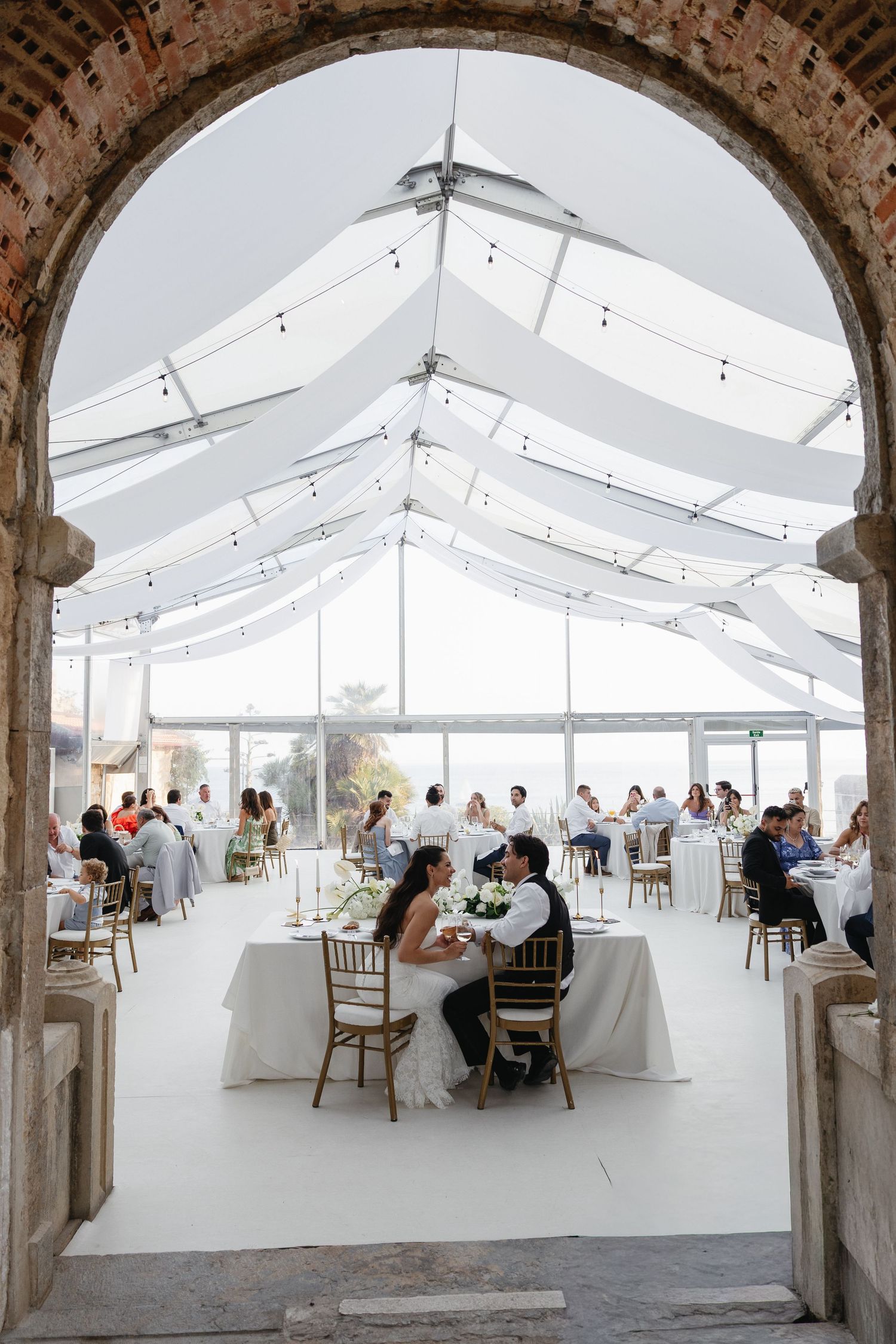 Elegant outdoor wedding reception under a clear tent with string lights, guests seated at white-draped tables, framed by a stone arch.