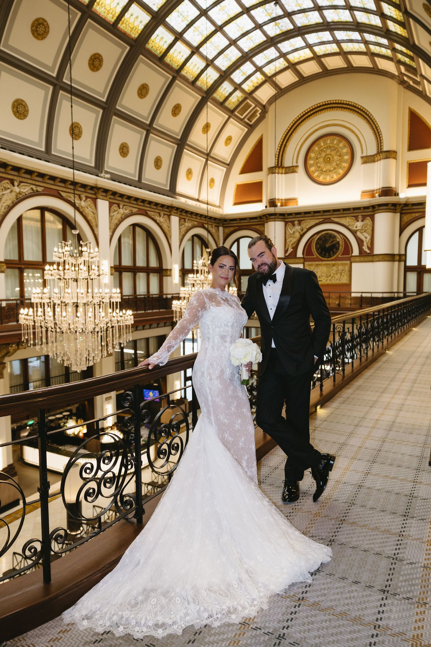 Bride in lace mermaid gown and groom in tuxedo pose on ornate balcony of elegant wedding venue with chandeliers.