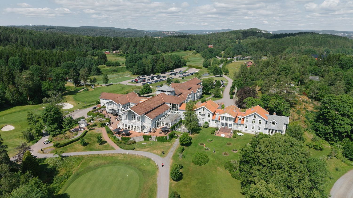 Aerial view of a golf resort with lush green fairways, red-roofed buildings, and surrounding dense forest landscape.