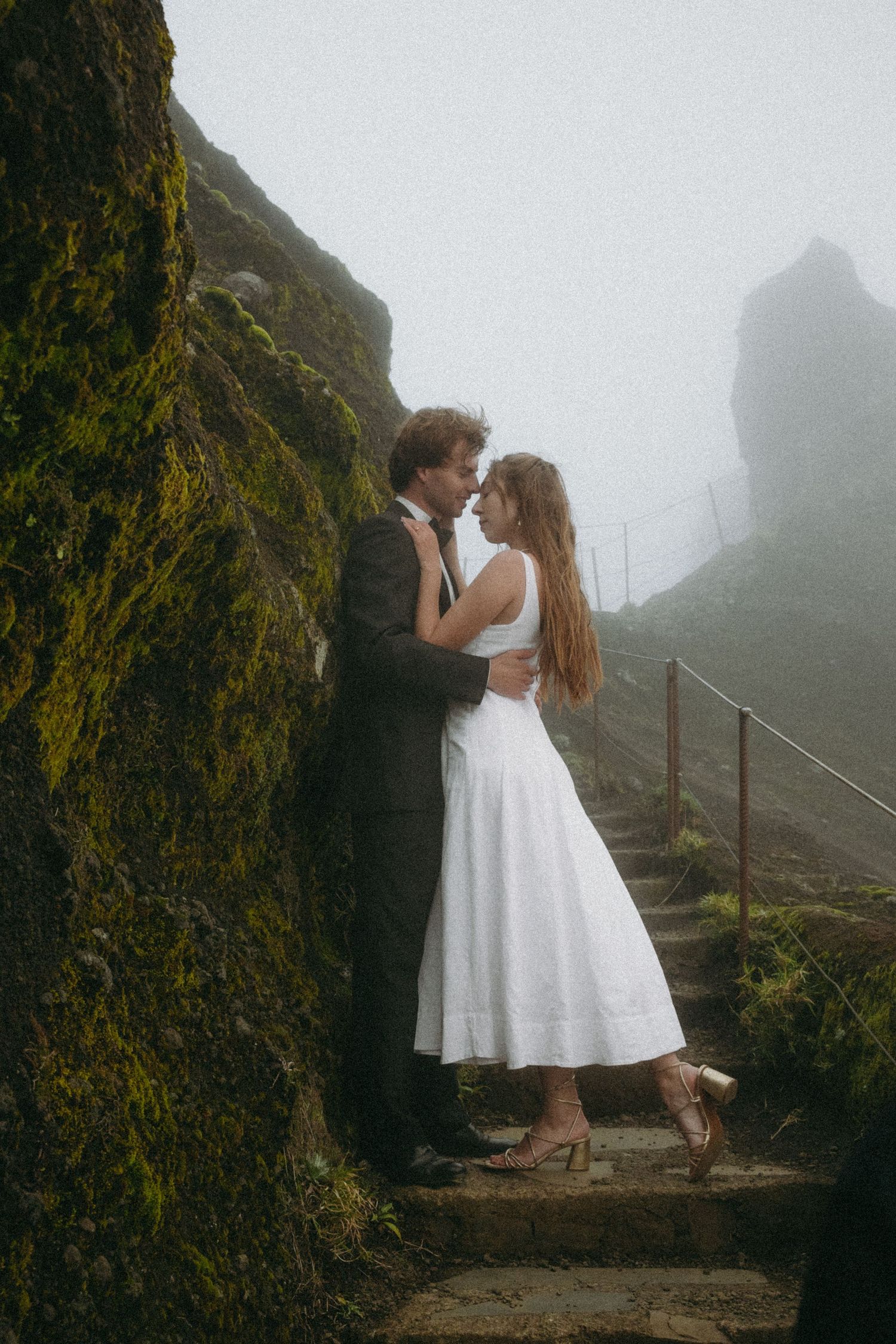 Bride and groom sharing romantic moment on misty mountain steps beside moss-covered cliff.