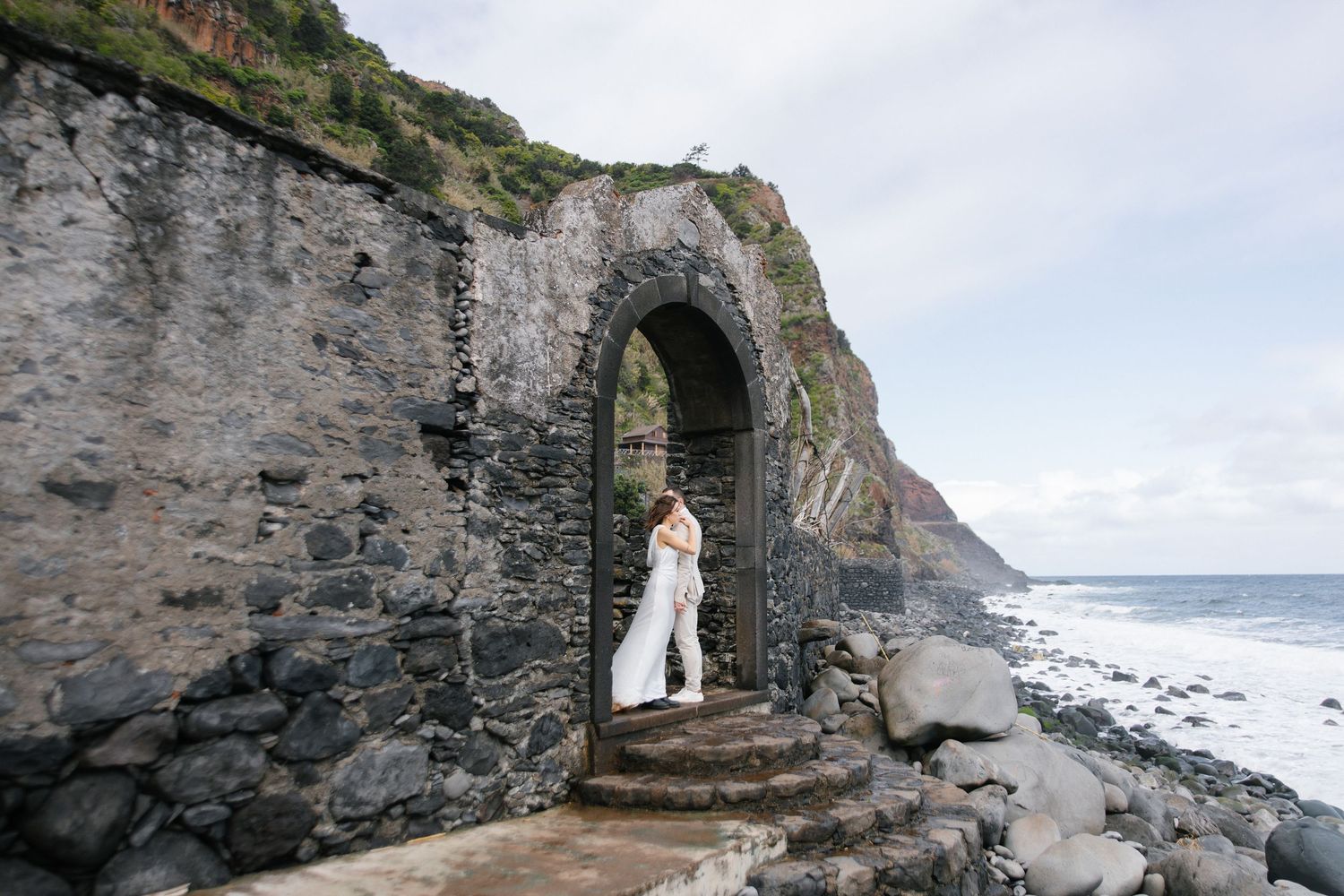Wedding couple embracing under a stone arch at a rocky coastal location in Madeira with ocean waves behind them.