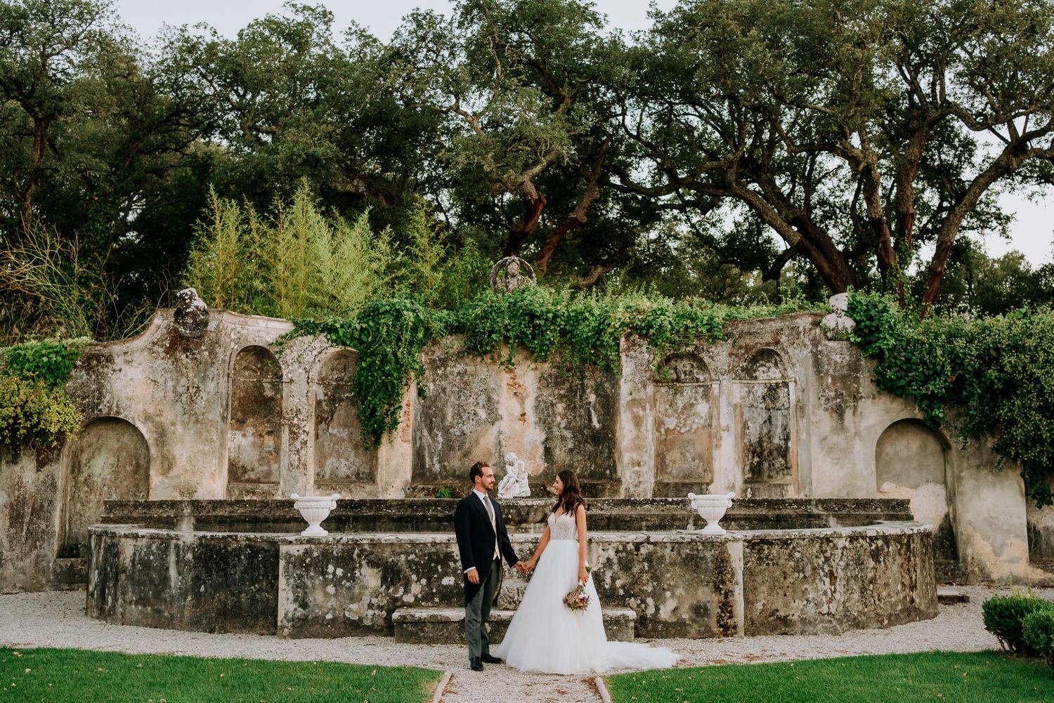 Newlyweds walking hand-in-hand in front of a moss-covered stone wall with arched niches at a garden wedding.