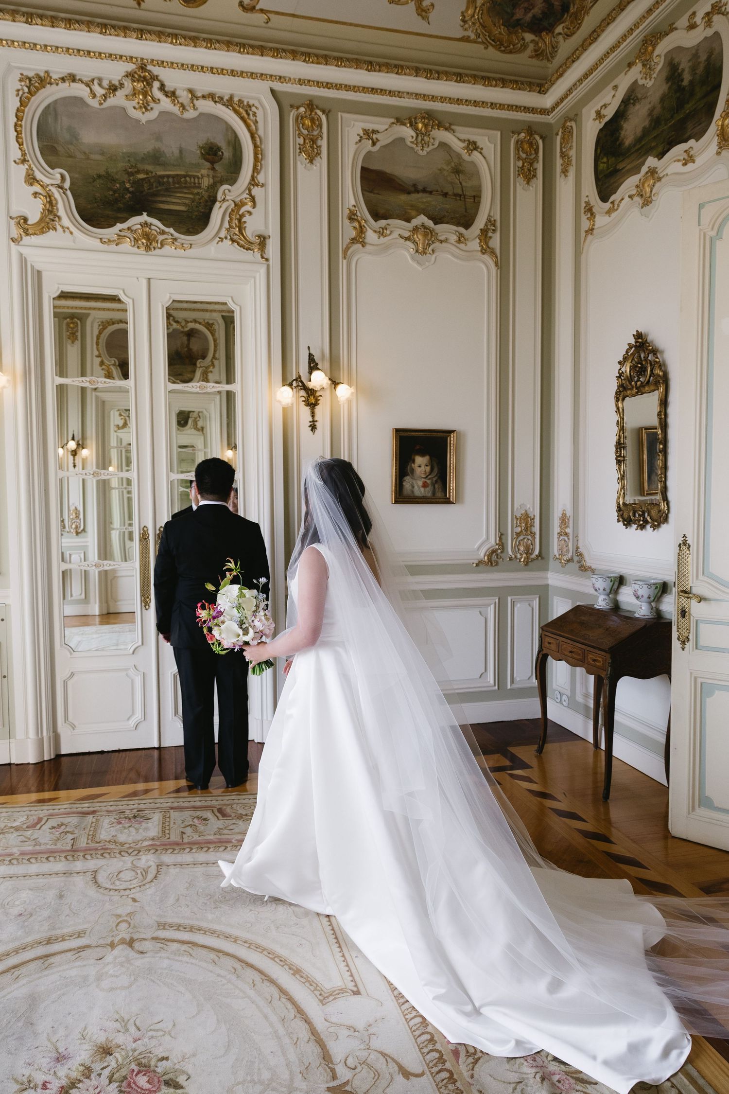 Bride in white gown with veil and groom in tuxedo stand in ornate Baroque ballroom with gold moldings and paintings.