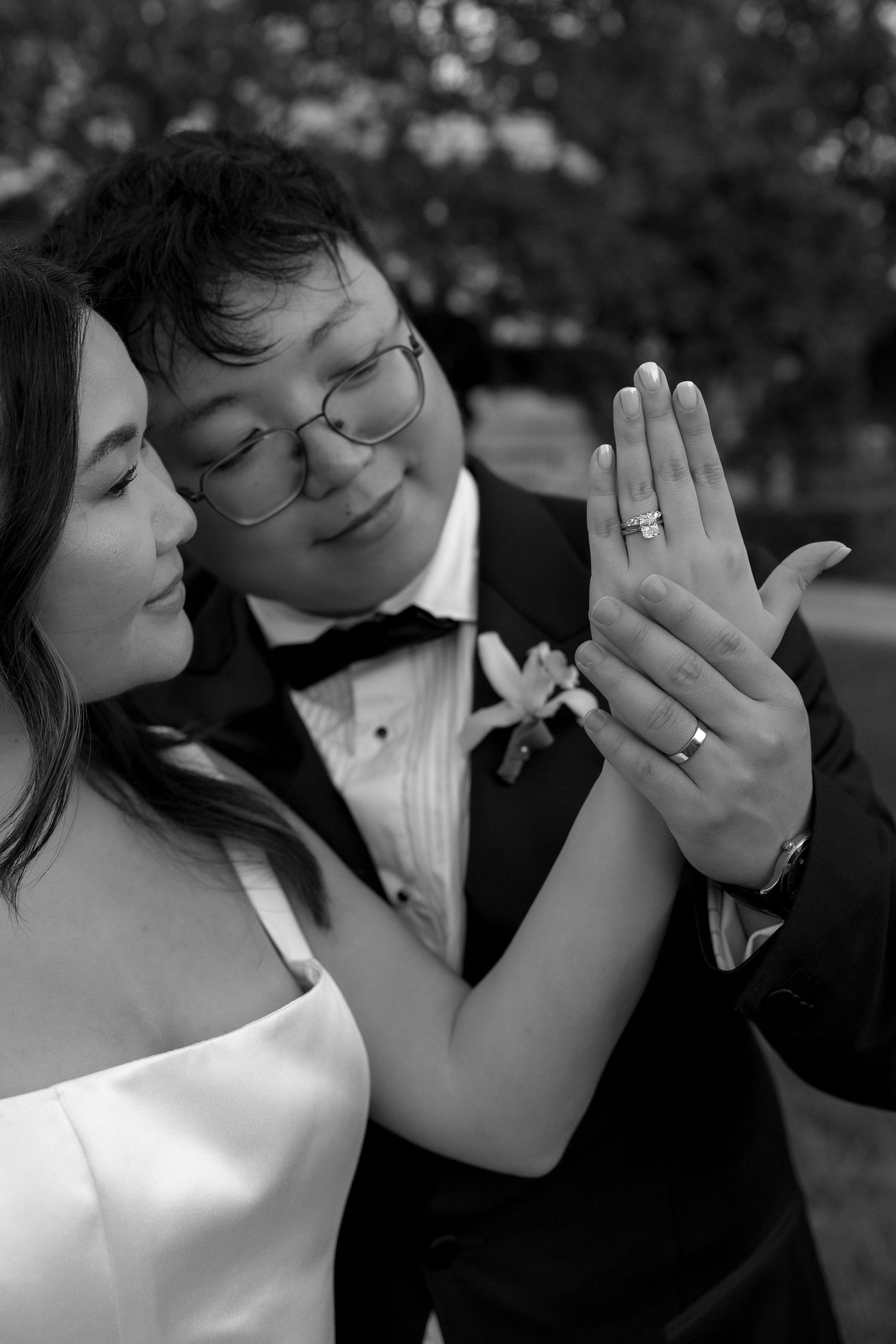 Black and white photo of newlyweds displaying wedding rings, groom in tuxedo with boutonniere, bride in strapless gown.