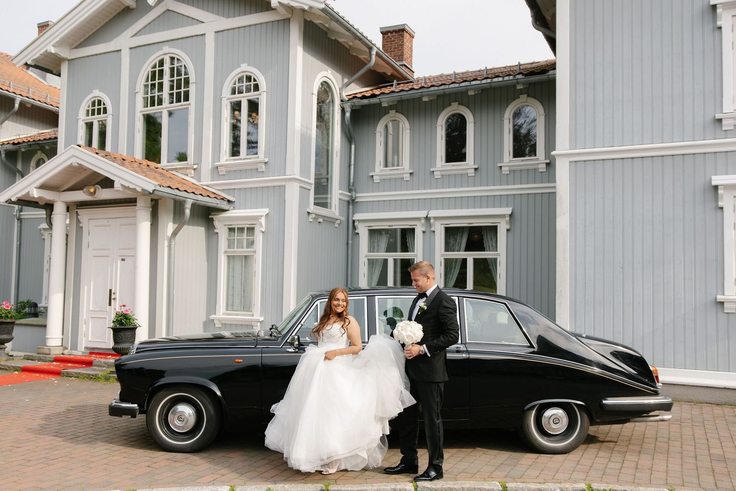 Bride and groom posing by a vintage black car in front of a elegant gray wooden building on their wedding day.