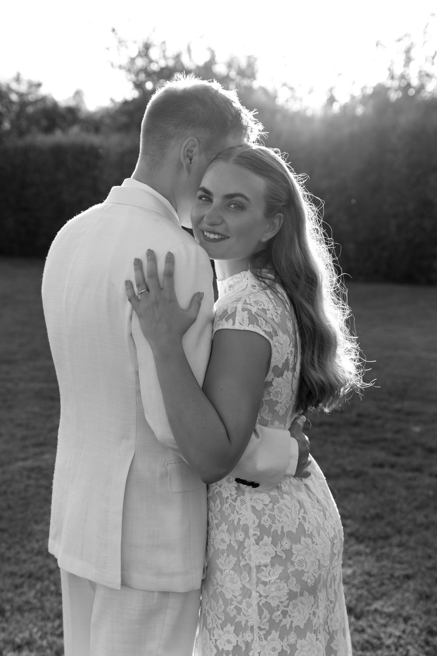 Black and white photo of a smiling bride embracing her groom outdoors at sunset.