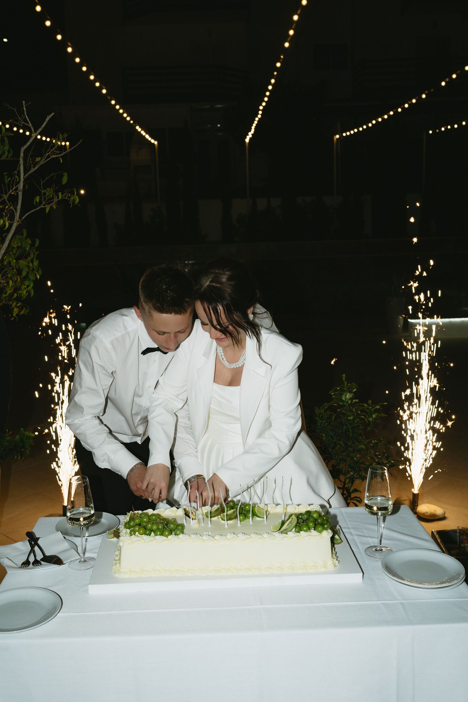 Bride and groom cutting white wedding cake decorated with green grapes outdoors at night with sparklers.