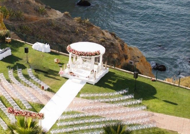Aerial view of stunning outdoor wedding ceremony setup on clifftop with floral arch and white chairs by the sea.