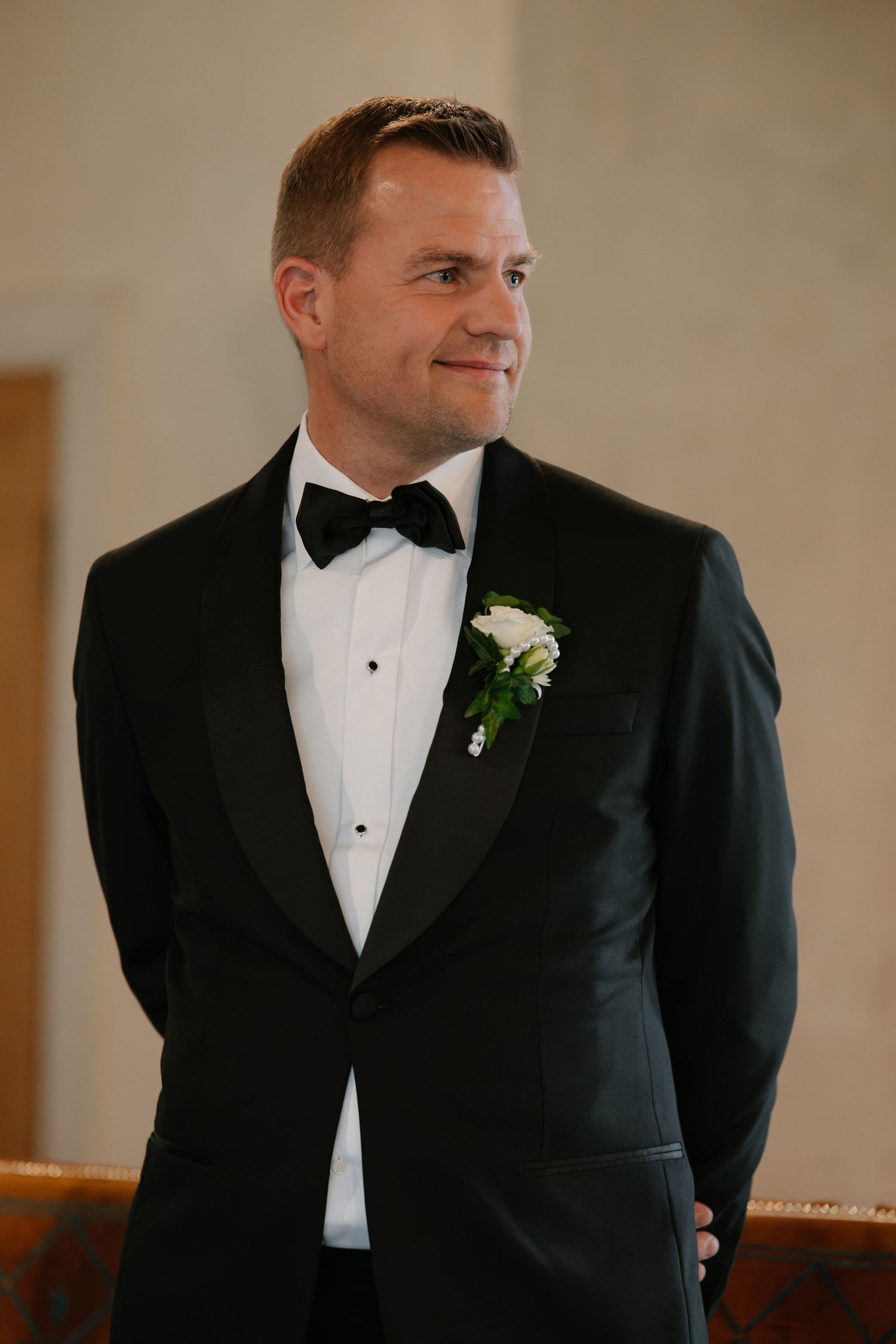 Groom in black tuxedo with white boutonniere and bow tie smiling at wedding ceremony.