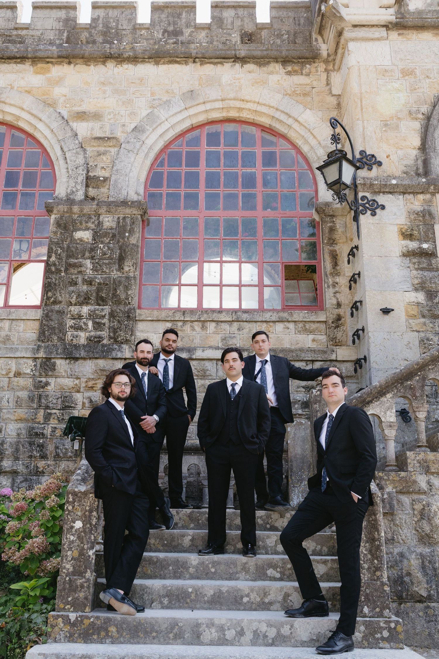 Six men in black suits posing on stone steps outside a historic building with arched red windows.