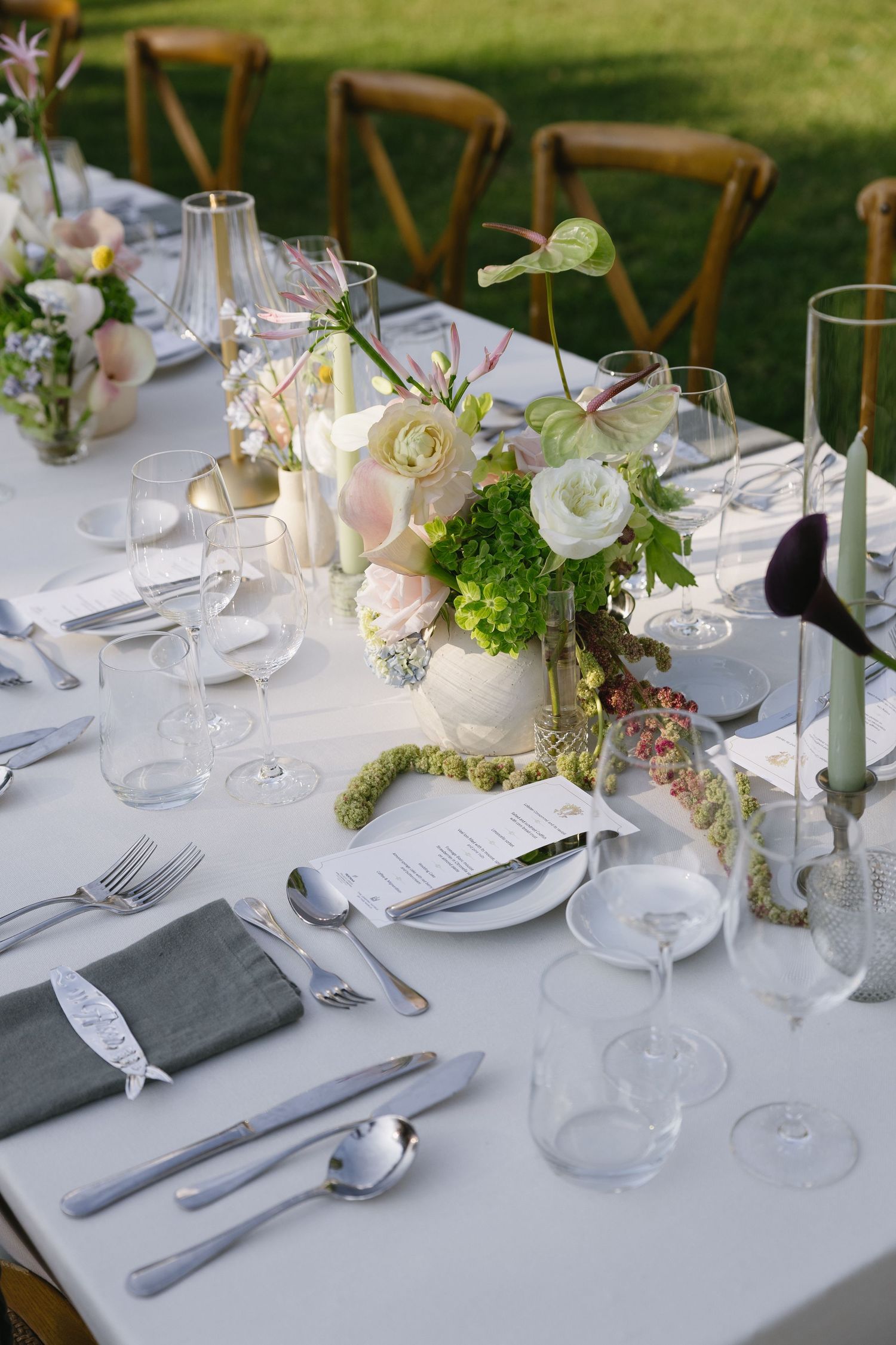 Elegant outdoor wedding reception table set with floral centerpieces, crystal glasses, gray napkins, and silverware on white linen.