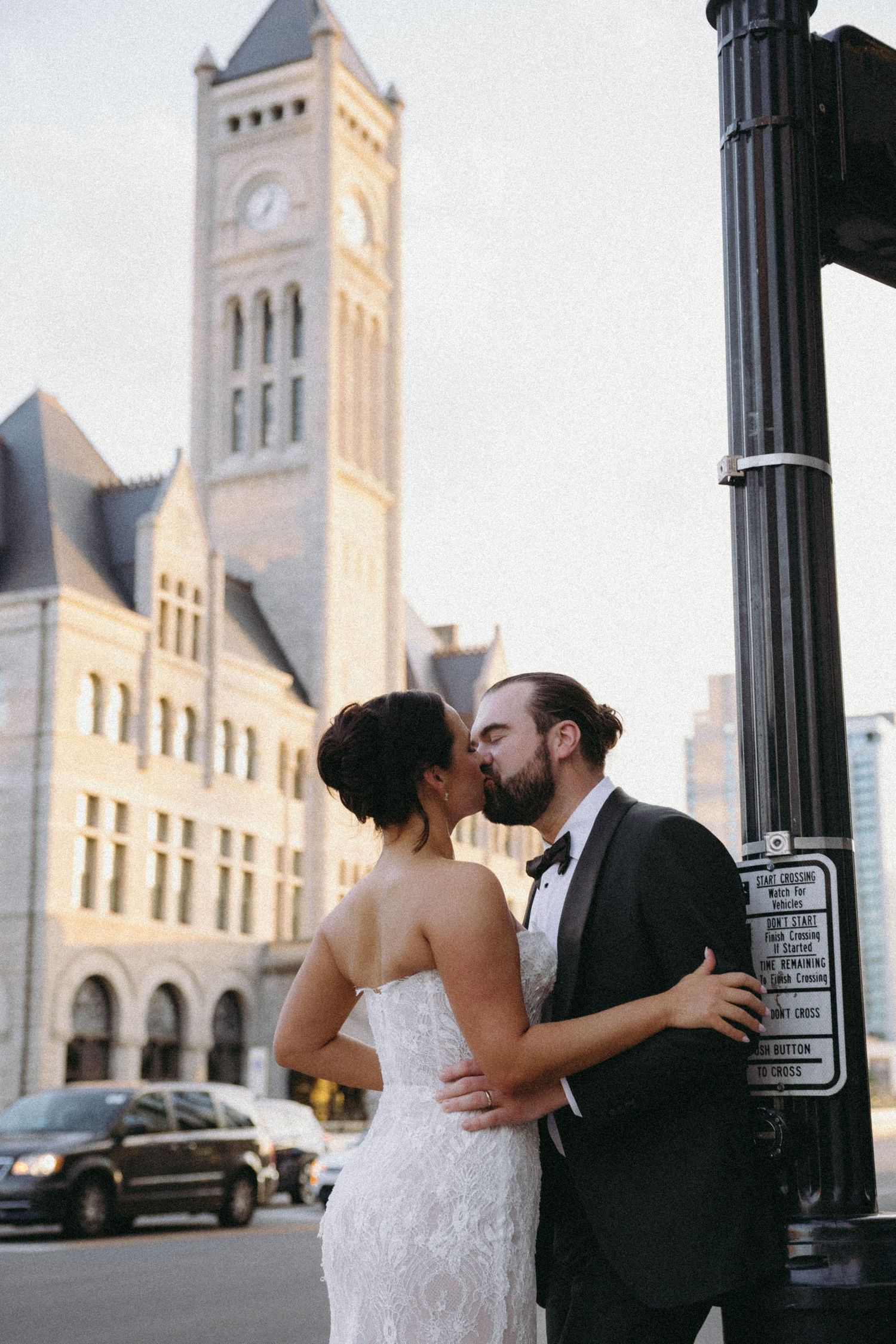 Bride and groom share a romantic kiss near a street lamp with a historic stone clock tower building in the background.