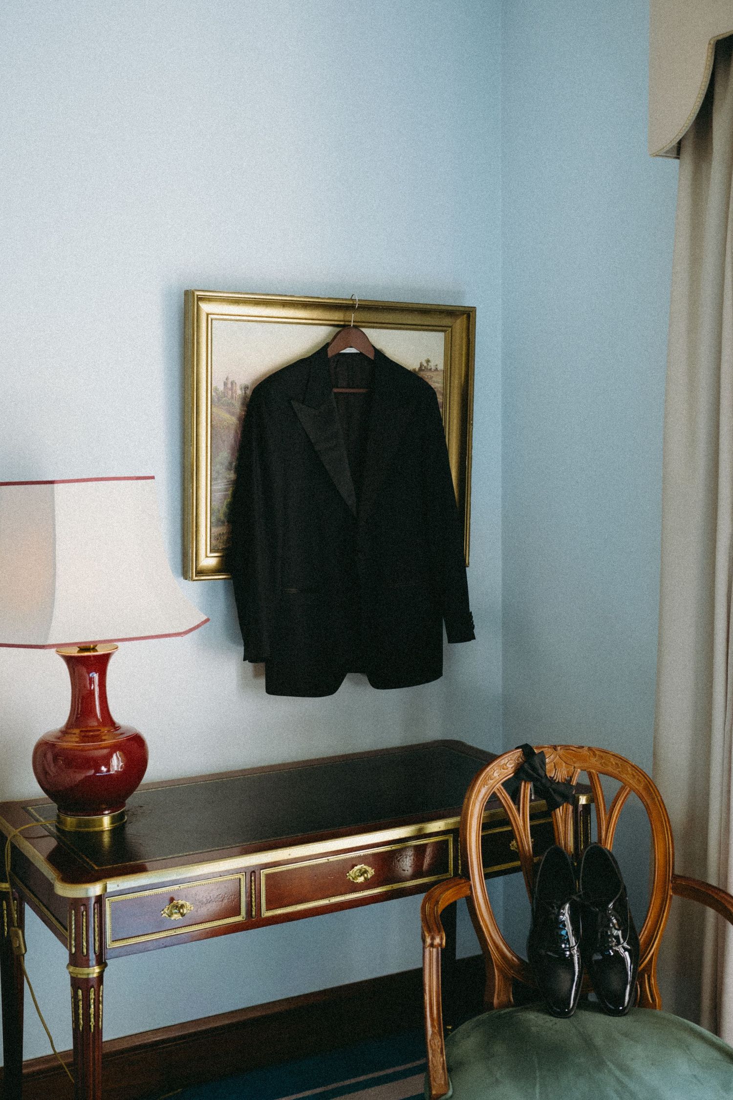 Black suit jacket on hanger over gold-framed art, beside red lamp on antique desk with dress shoes on wooden chair.