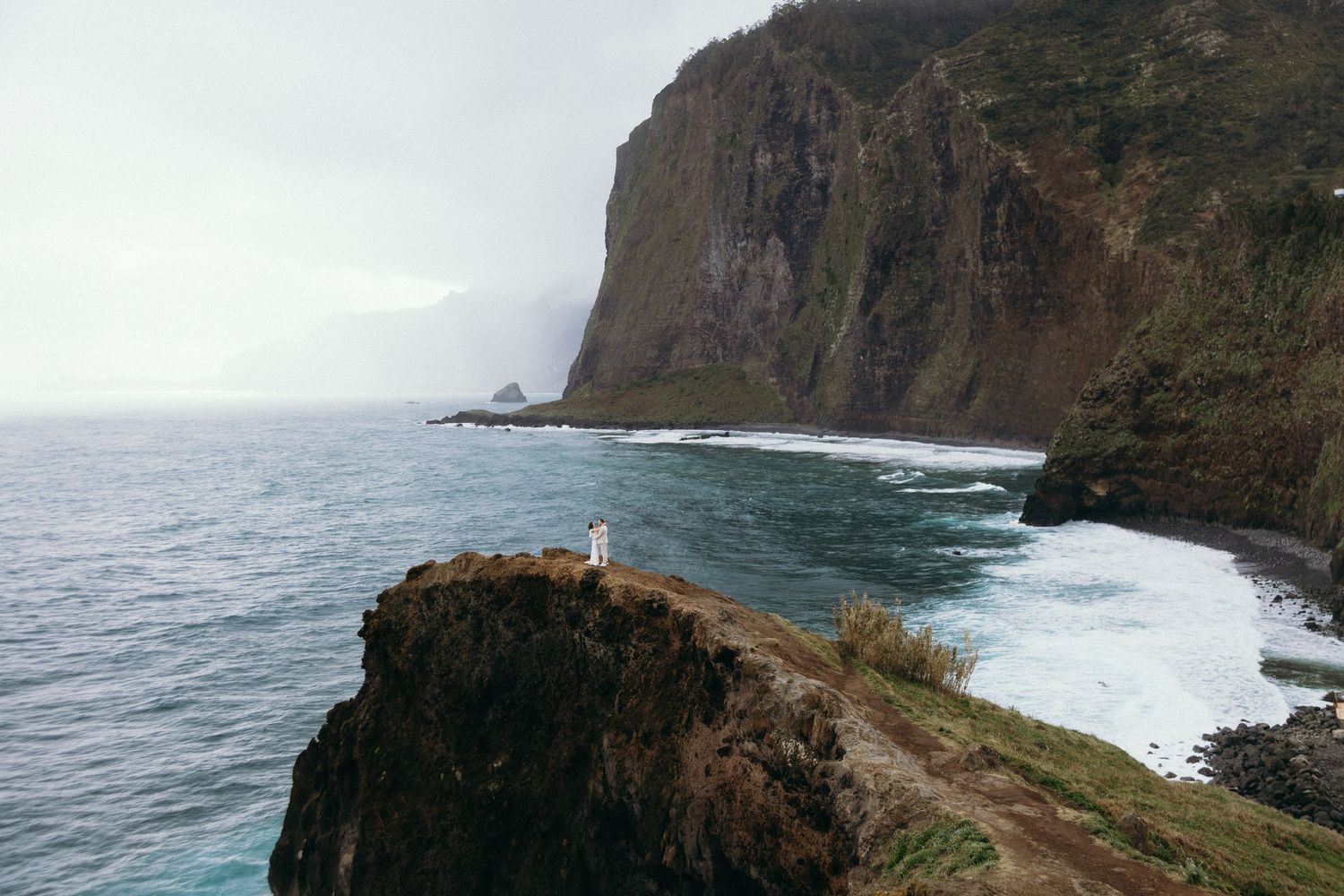 Lone figure in white dress stands on rocky coastal cliff overlooking crashing waves and dramatic cliffs.