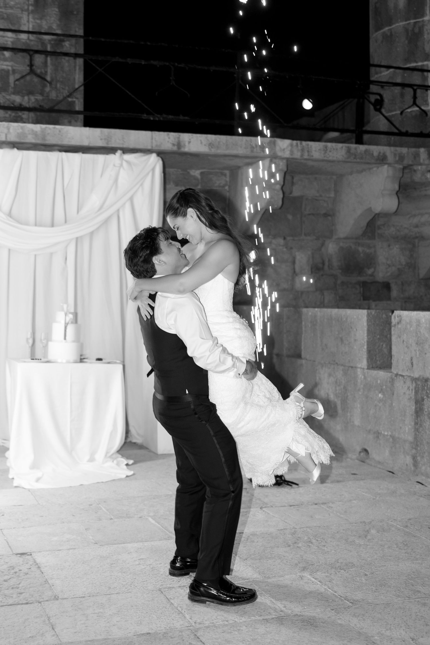 Black and white photo of groom lifting bride during first dance with sparkling lights at elegant wedding reception.