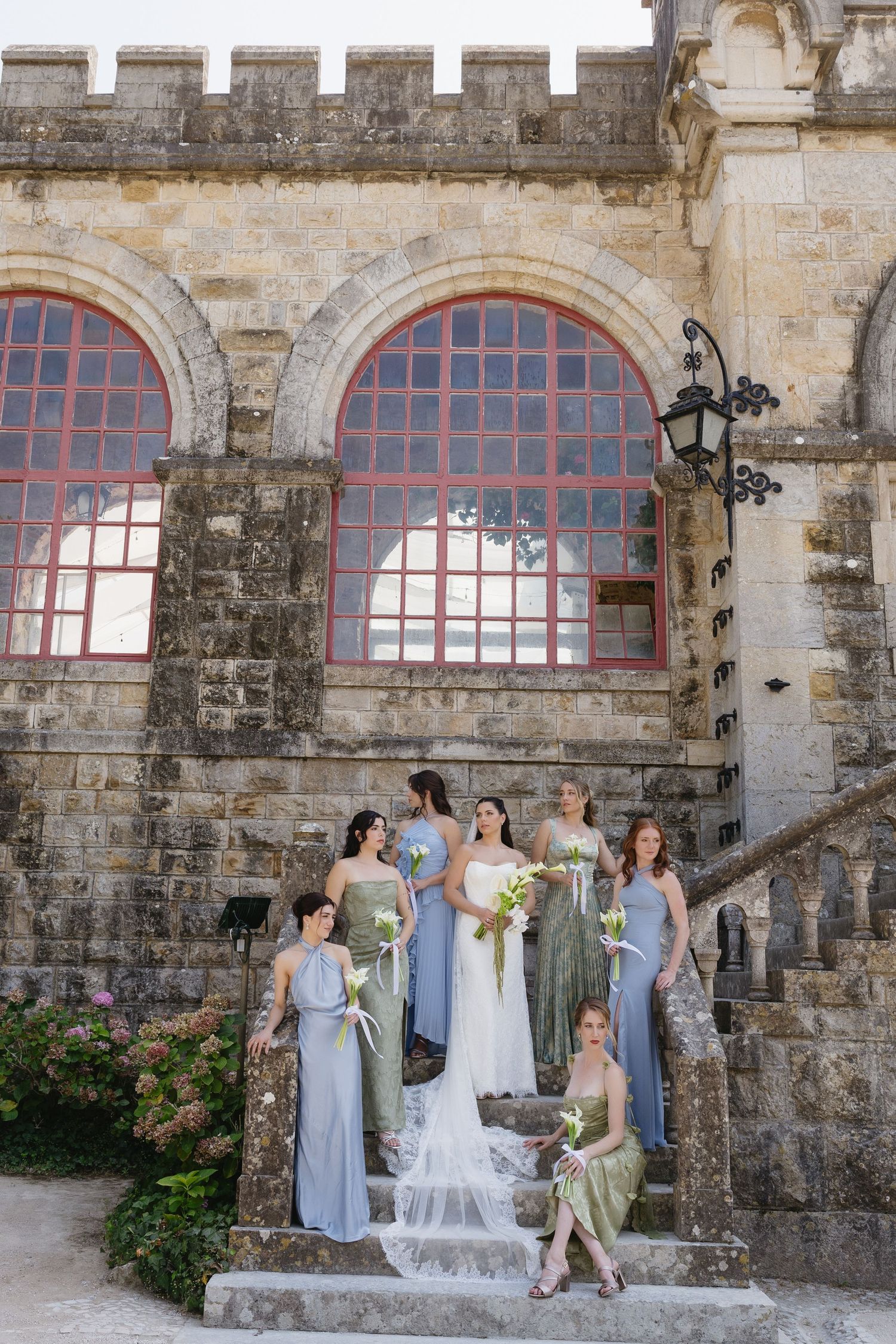 Bride and bridesmaids in blue gowns pose on stone steps of a castle venue with arched red windows.