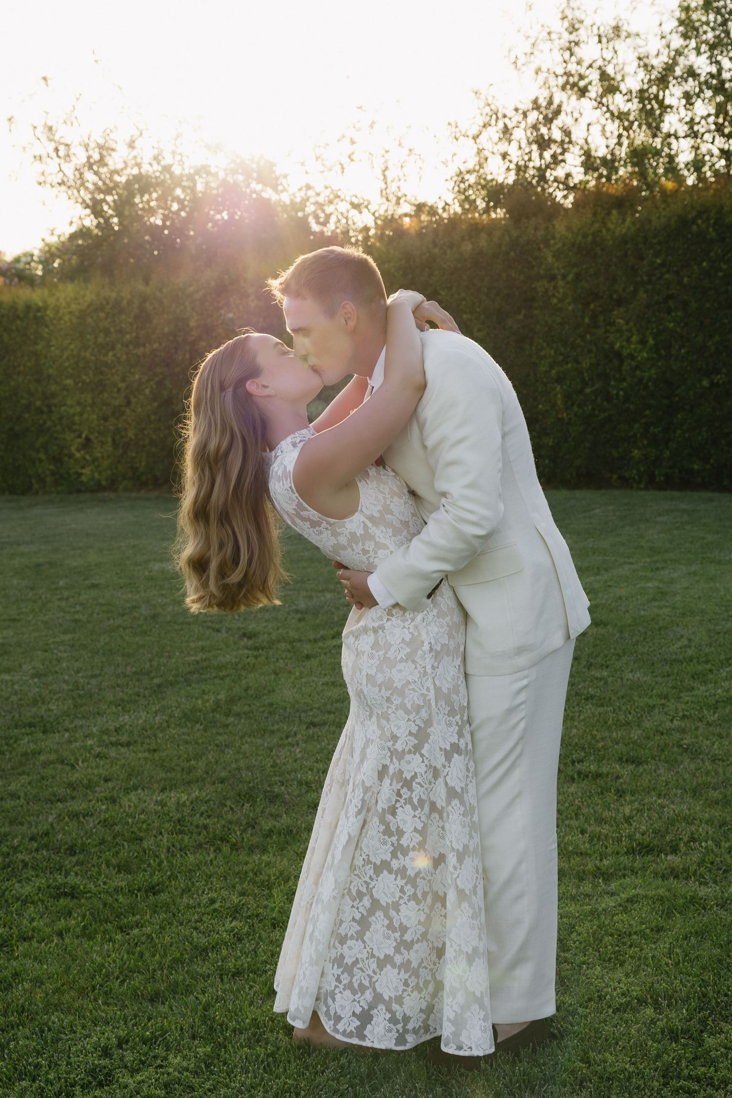 Bride and groom share a romantic sunset kiss on green lawn, bride in lace gown, groom in white suit.