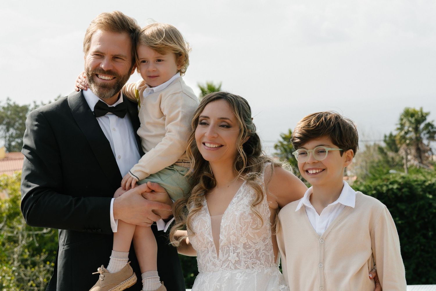 Smiling bride and groom in formal wear pose with two young children outdoors.