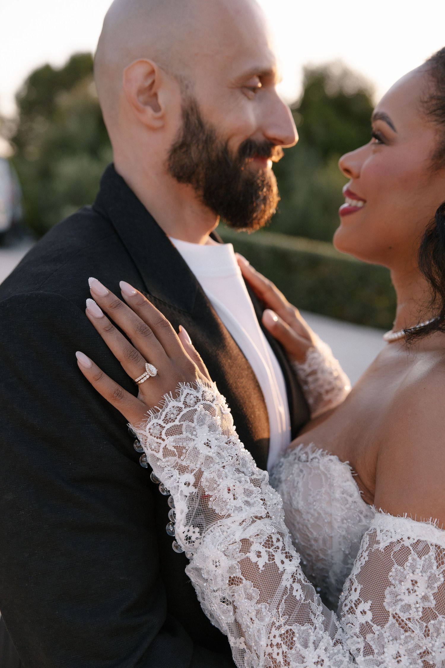 Close-up of bride in lace gown showing diamond engagement ring while embracing bearded groom at golden hour.