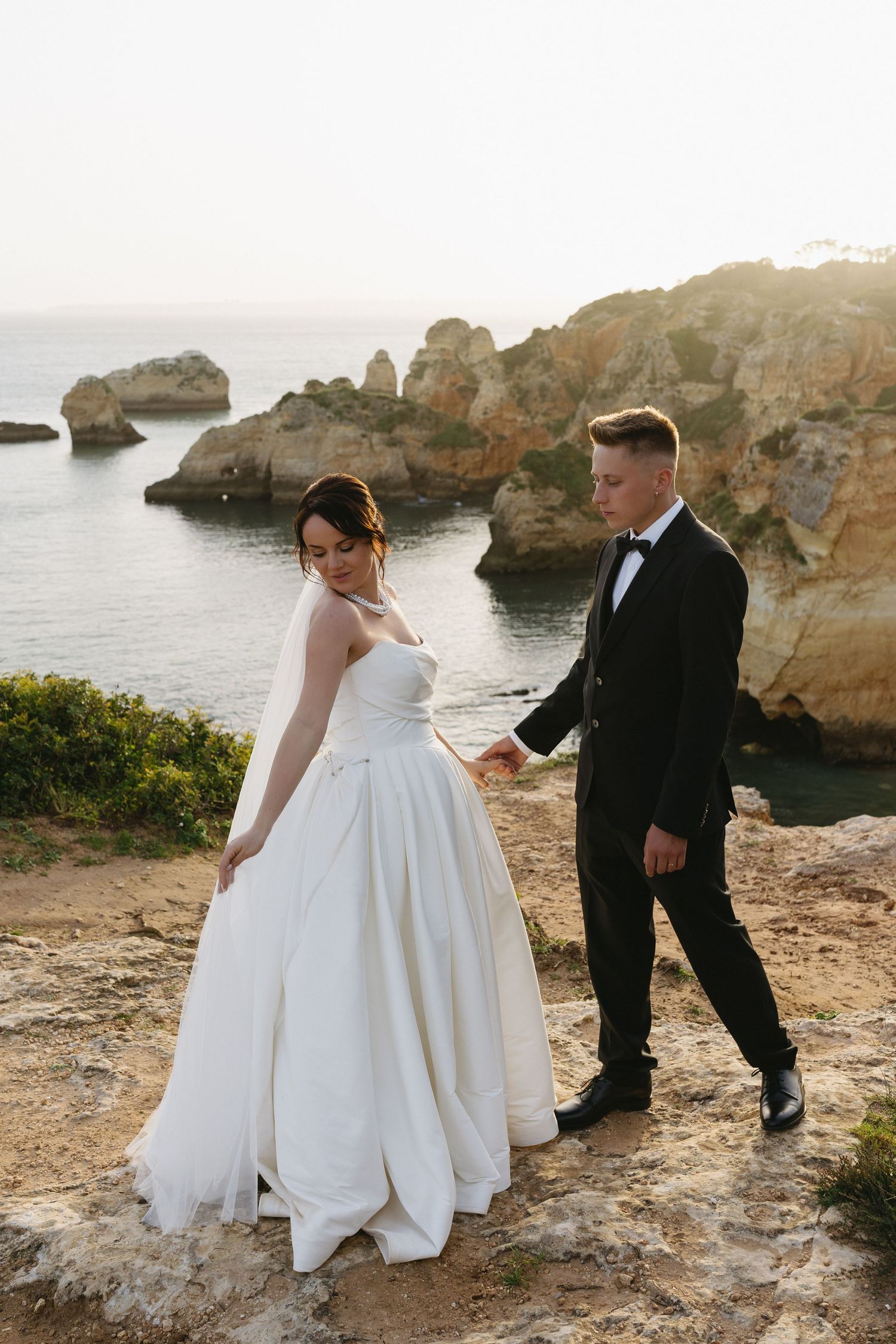 Bride and groom holding hands walking on rocky coastal cliffs at sunset with ocean views behind them.