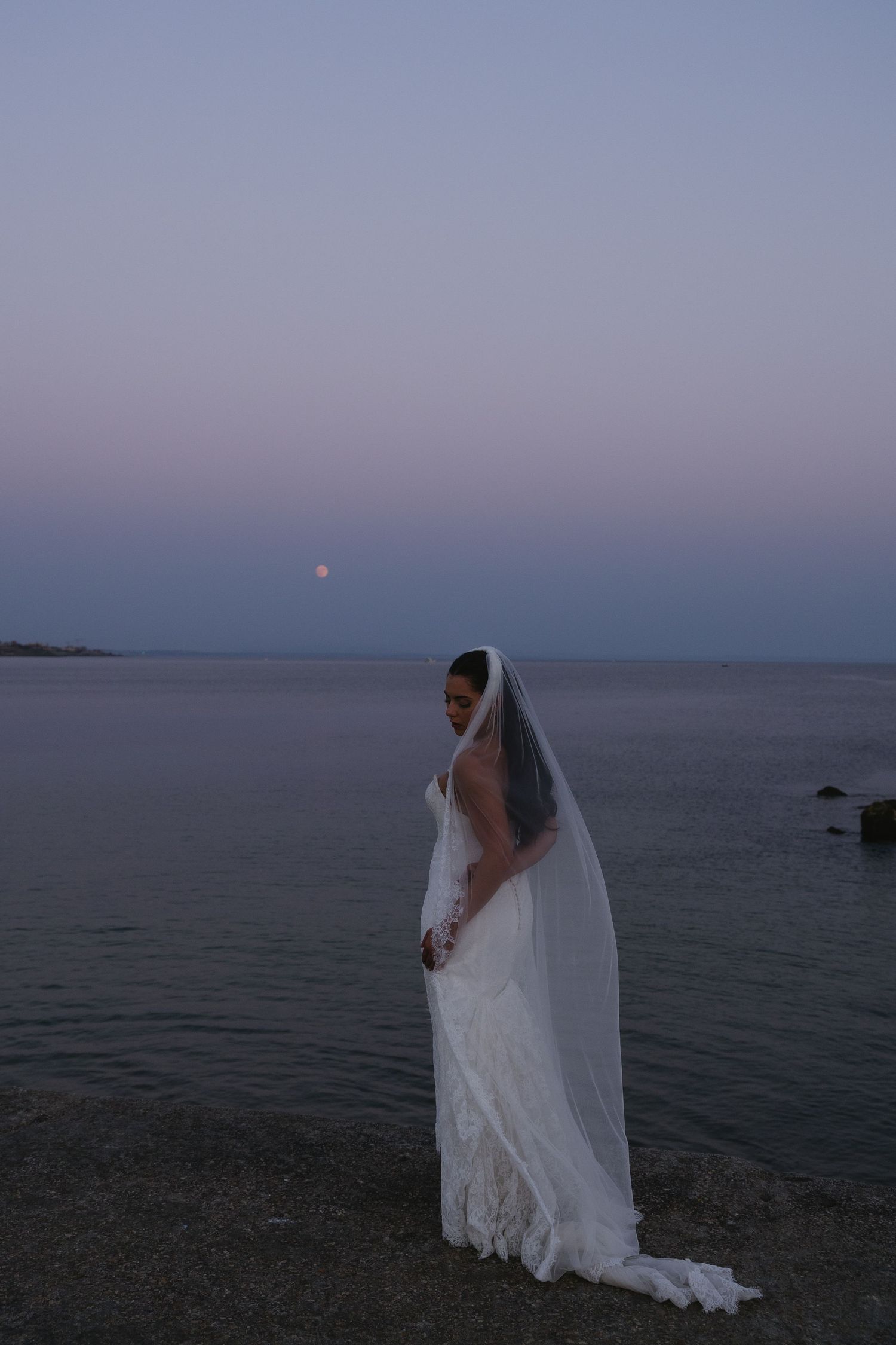 Bride in white gown and veil stands by the ocean shore at dusk with a full moon rising over the horizon.