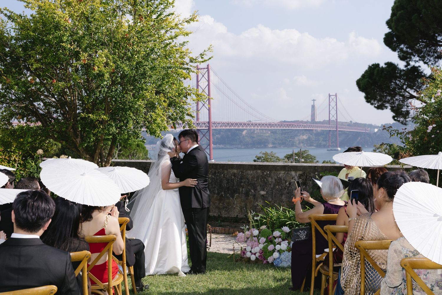 Outdoor wedding ceremony with couple kissing, guests holding white parasols, and the 25 de Abril Bridge over the Tagus River in Lisbon.