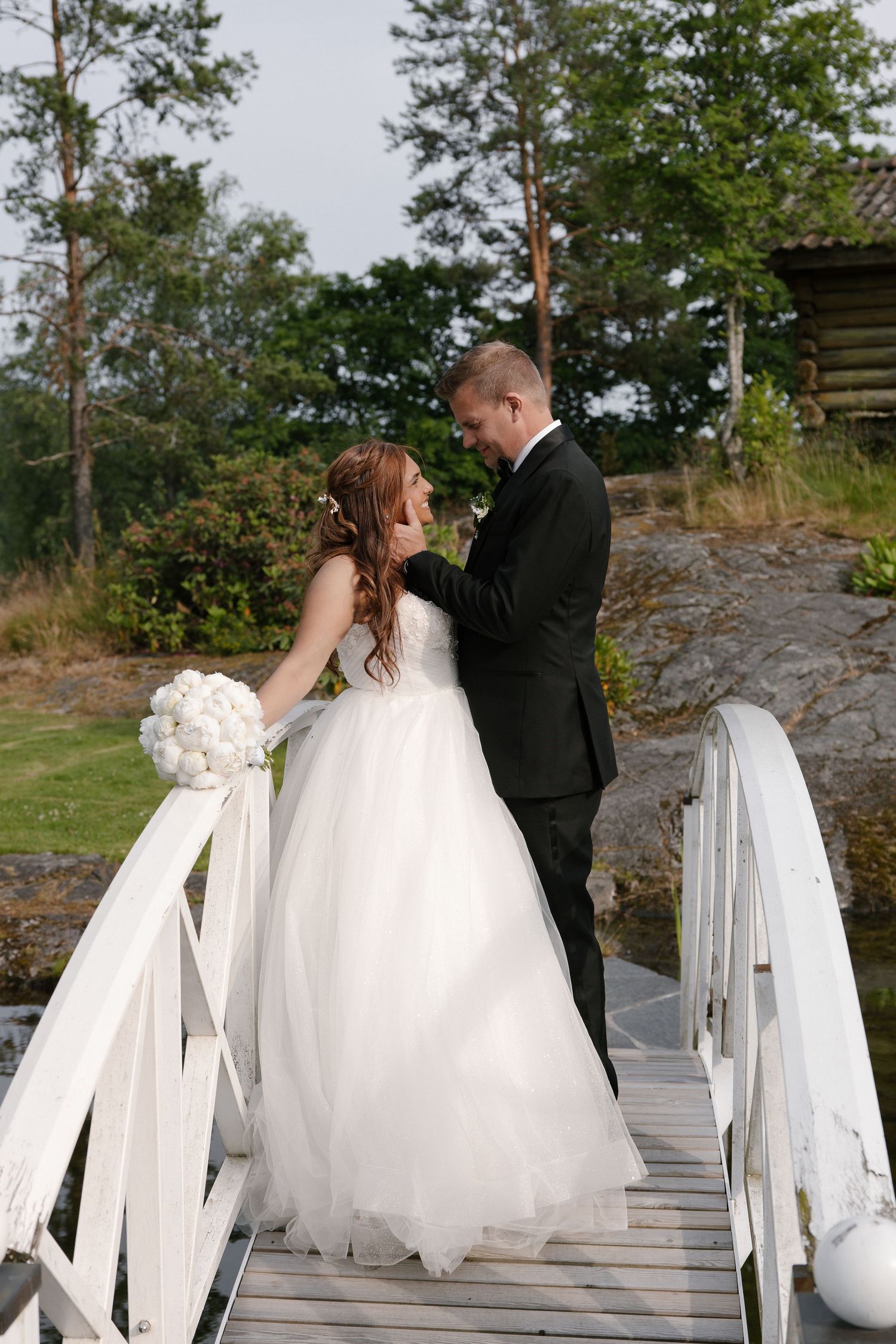 Bride and groom share a romantic moment on a white wooden bridge surrounded by lush greenery on their wedding day.
