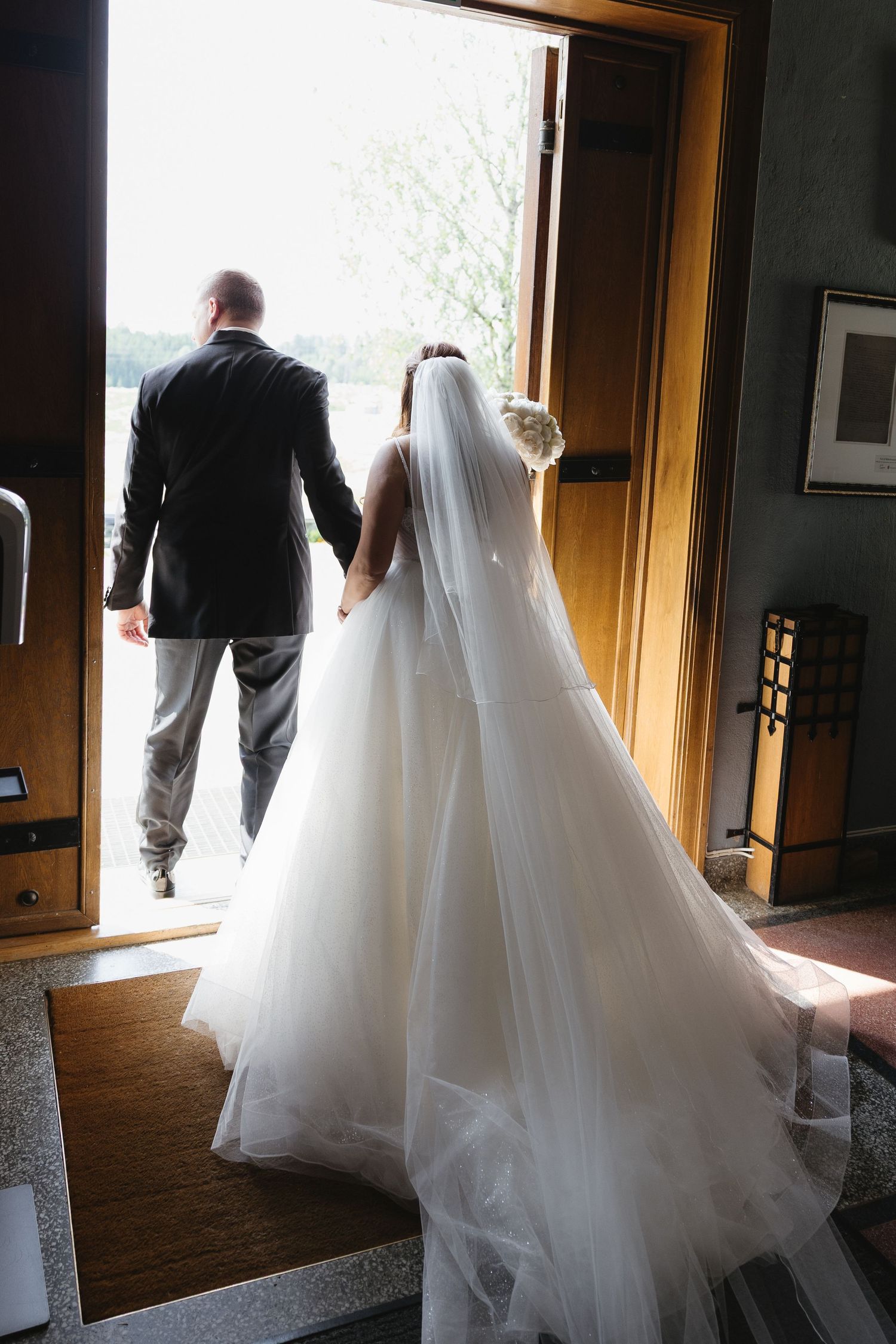 Bride in white gown and veil walks with groom through open doors at wedding venue.