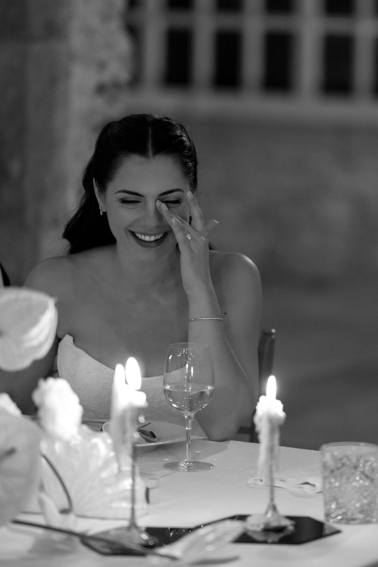 Black and white photo of a laughing bride wiping tears at a candlelit wedding reception table.