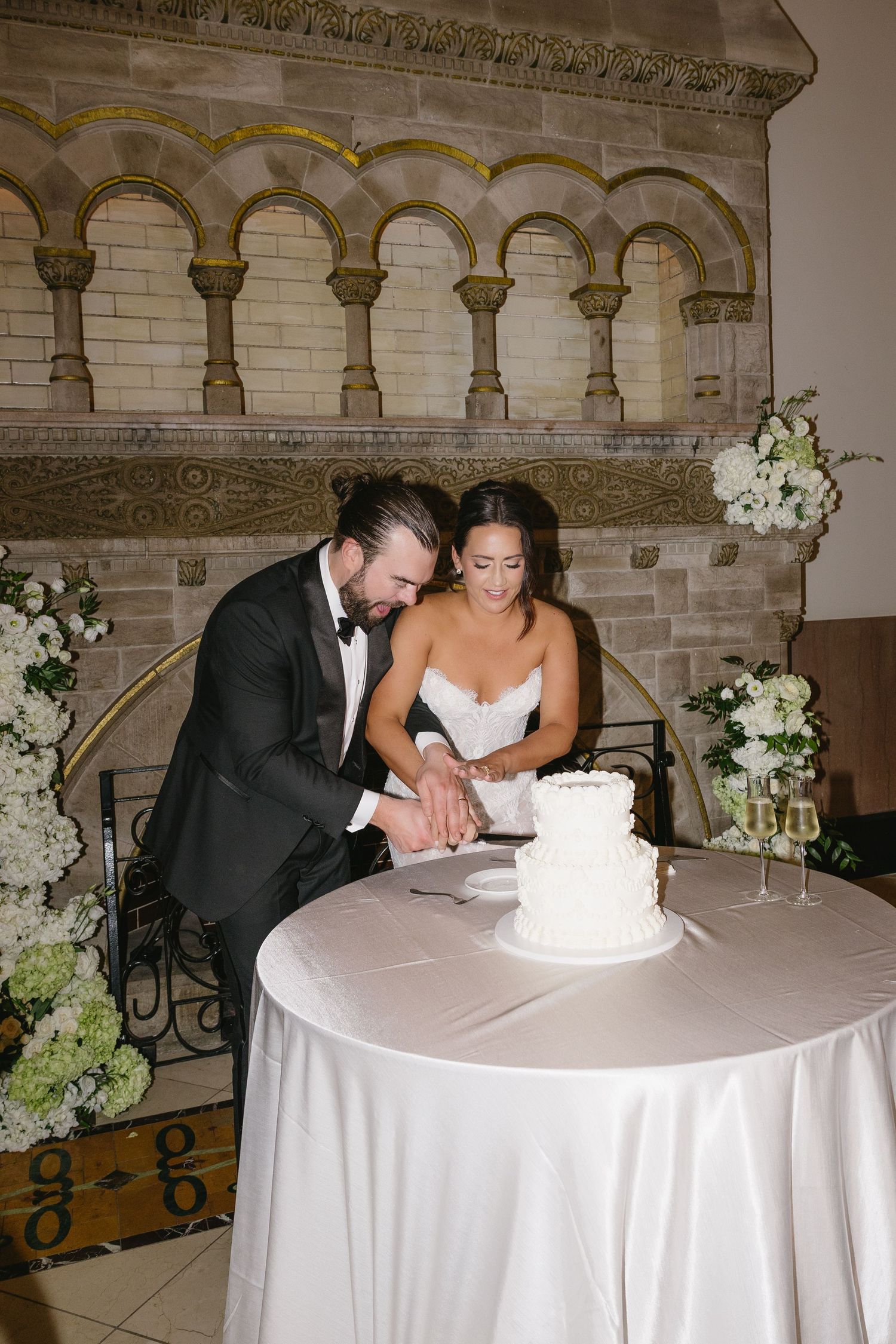 Bride and groom cutting white two-tier wedding cake at elegant reception venue with stone archway backdrop.