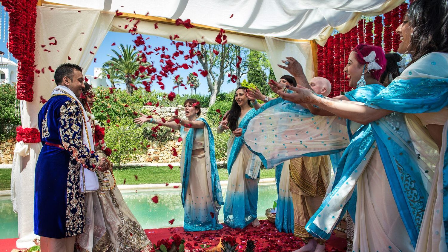 Colorful Indian wedding ceremony with rose petals shower, couple in traditional attire, and bridesmaids in blue outfits.