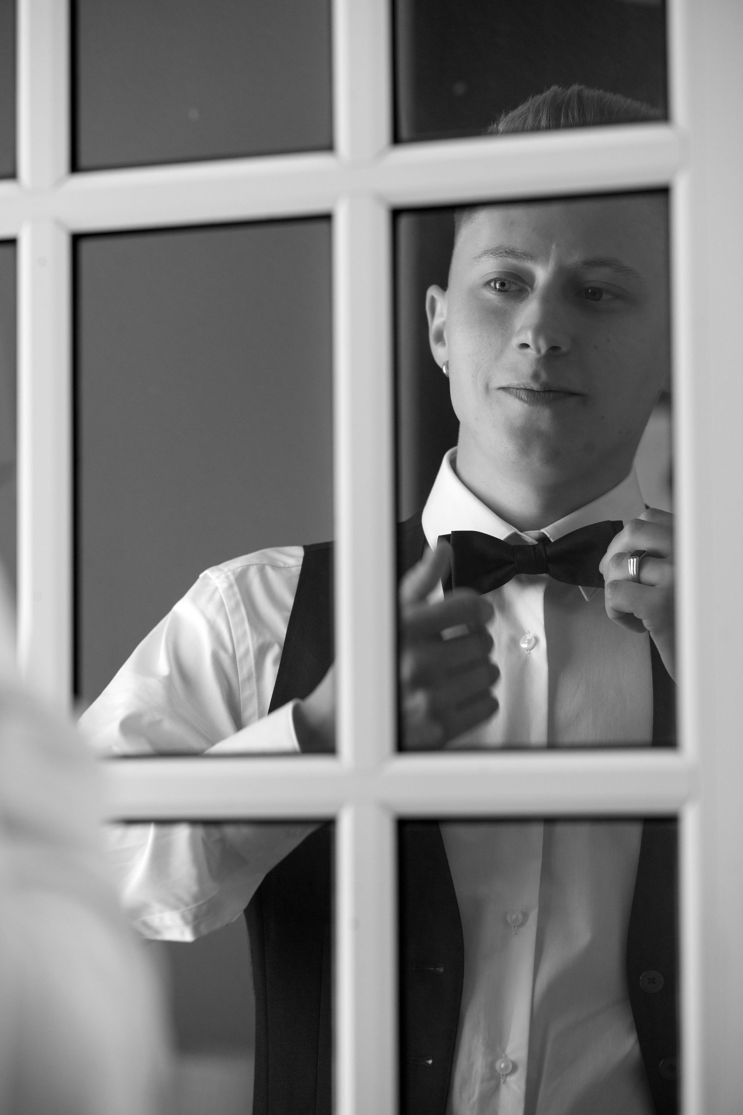 Black and white reflection of groom adjusting bow tie while looking into a French door mirror.
