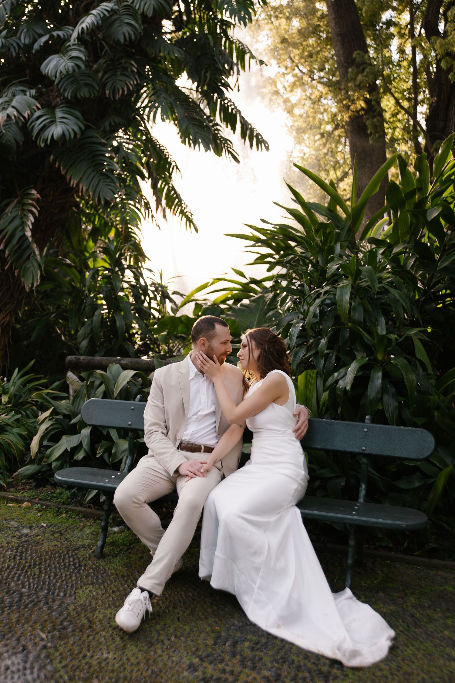 Couple sits intimately on a green bench surrounded by golden sunlit tropical garden greenery.