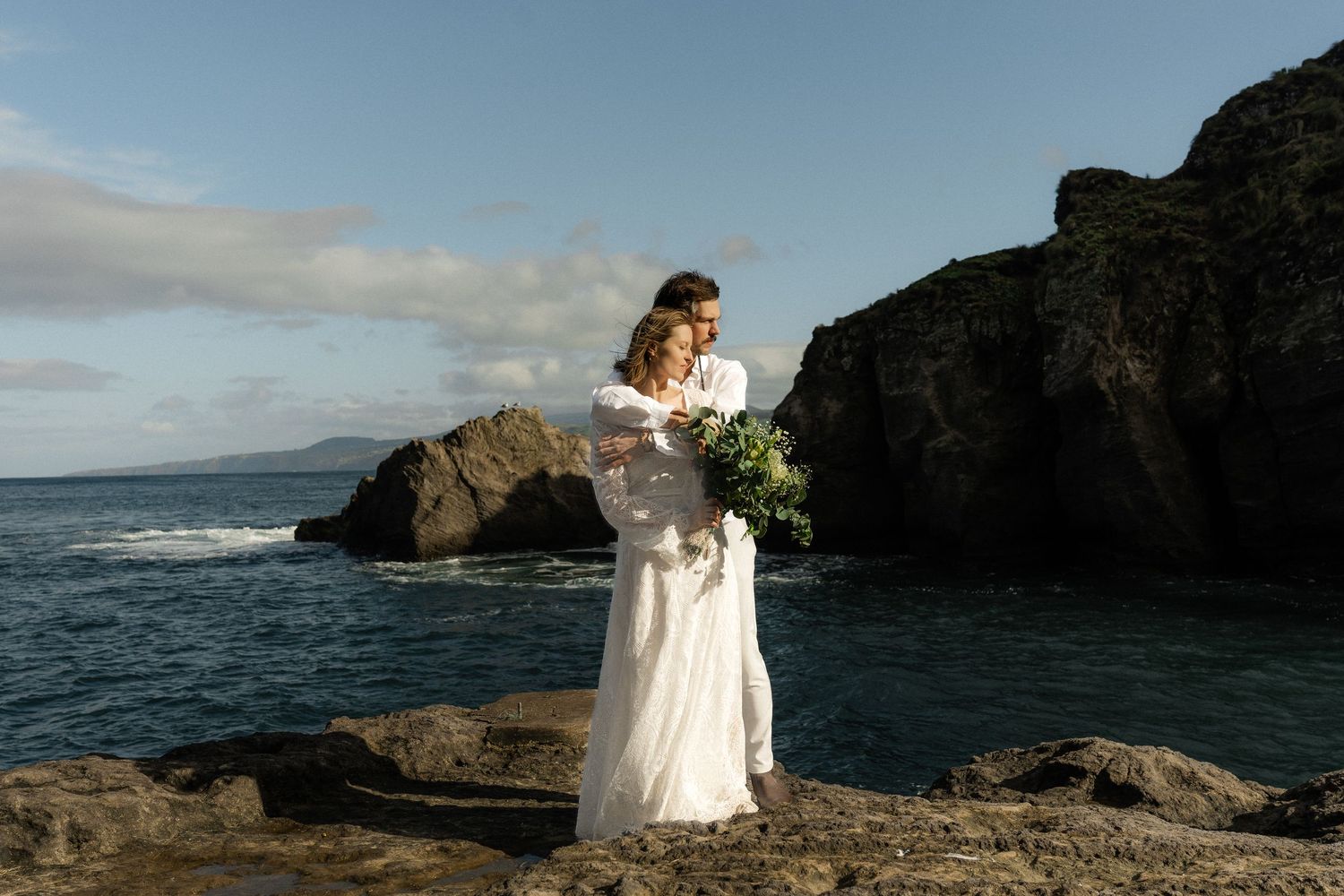 Bride in white lace gown holding bouquet standing on rocky coastal cliffs with ocean and sea stacks behind.