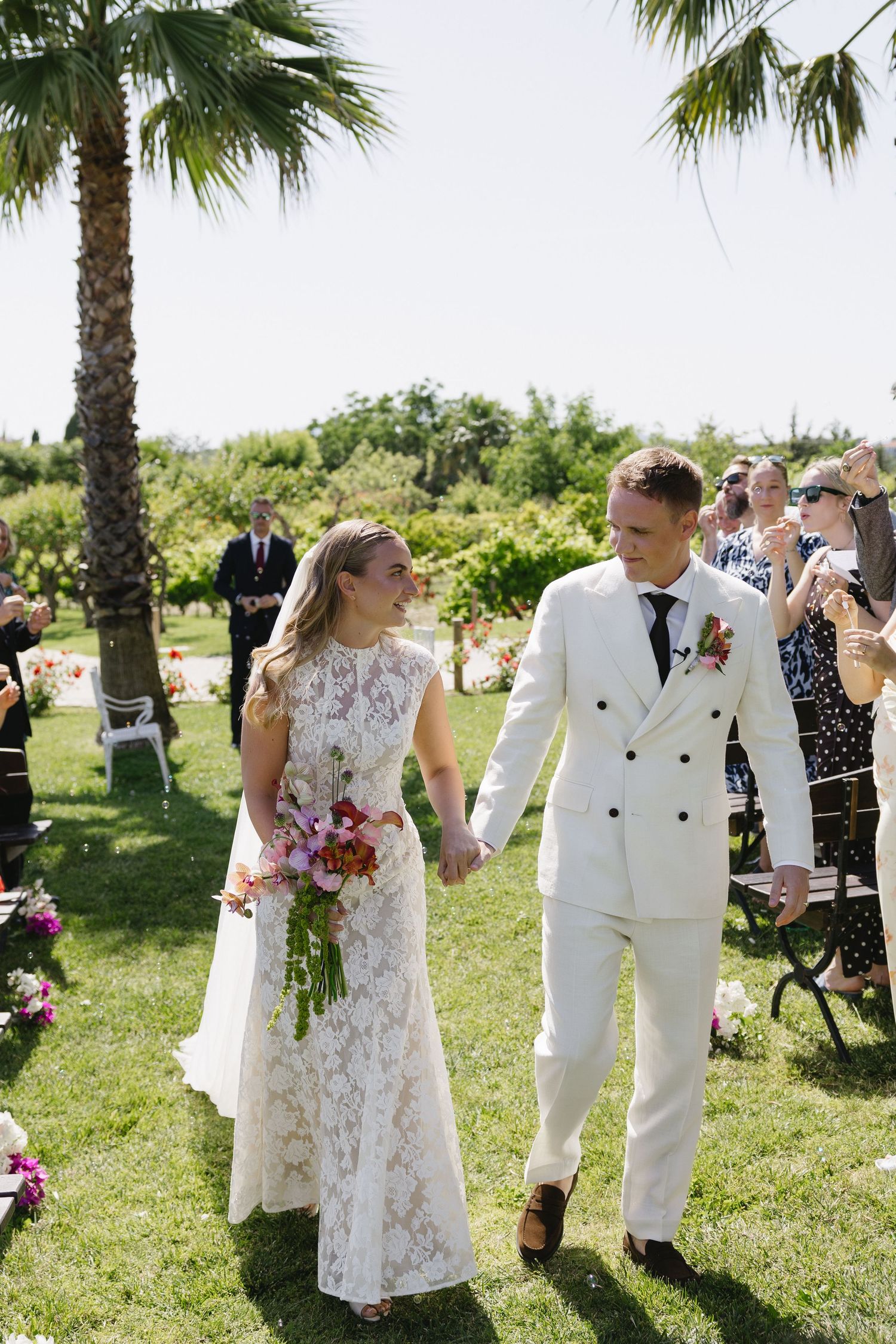 Newlyweds walk down grassy aisle hand-in-hand, bride in lace gown holding colorful bouquet, guests applauding.
