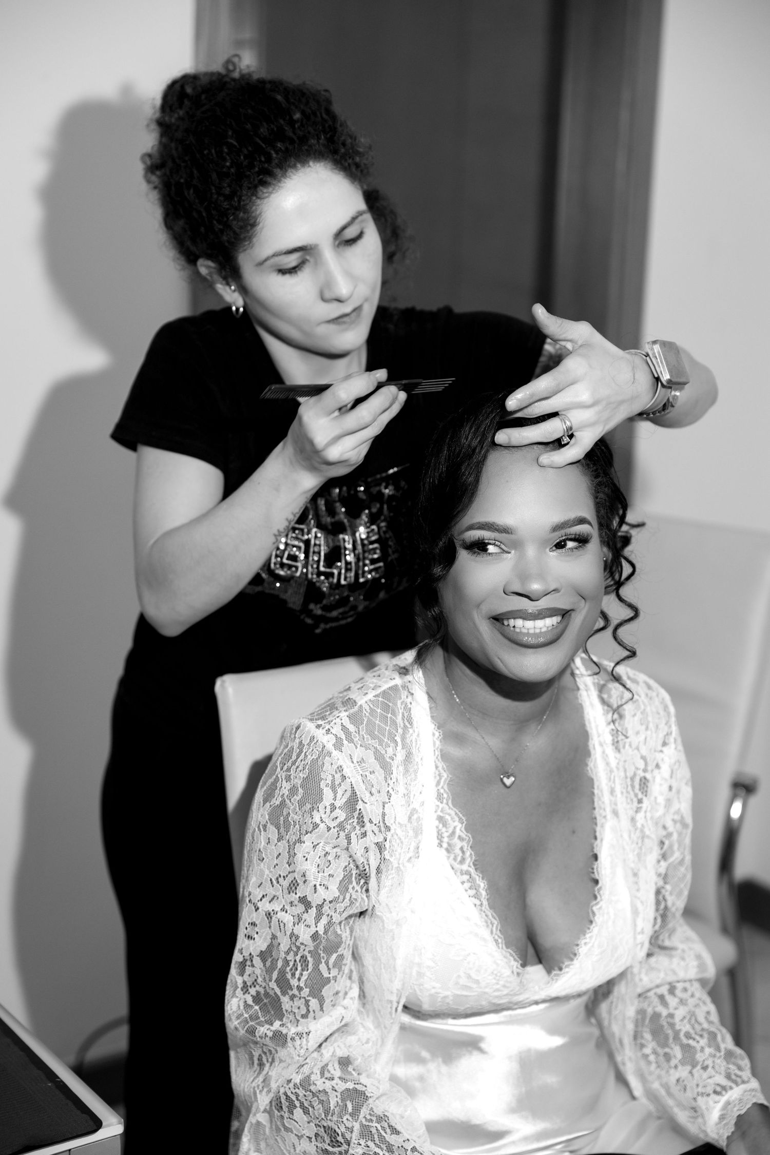 Black and white photo of makeup artist styling smiling bride's hair before wedding ceremony indoors.