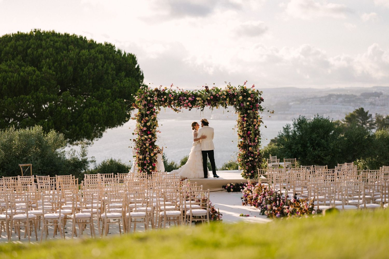 Bride and groom stand under floral arch at outdoor wedding ceremony with ocean view and elegant gold chairs.