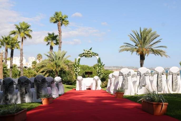 Outdoor wedding ceremony setup with red carpet aisle, white chair covers, floral arch, and tropical palm trees.