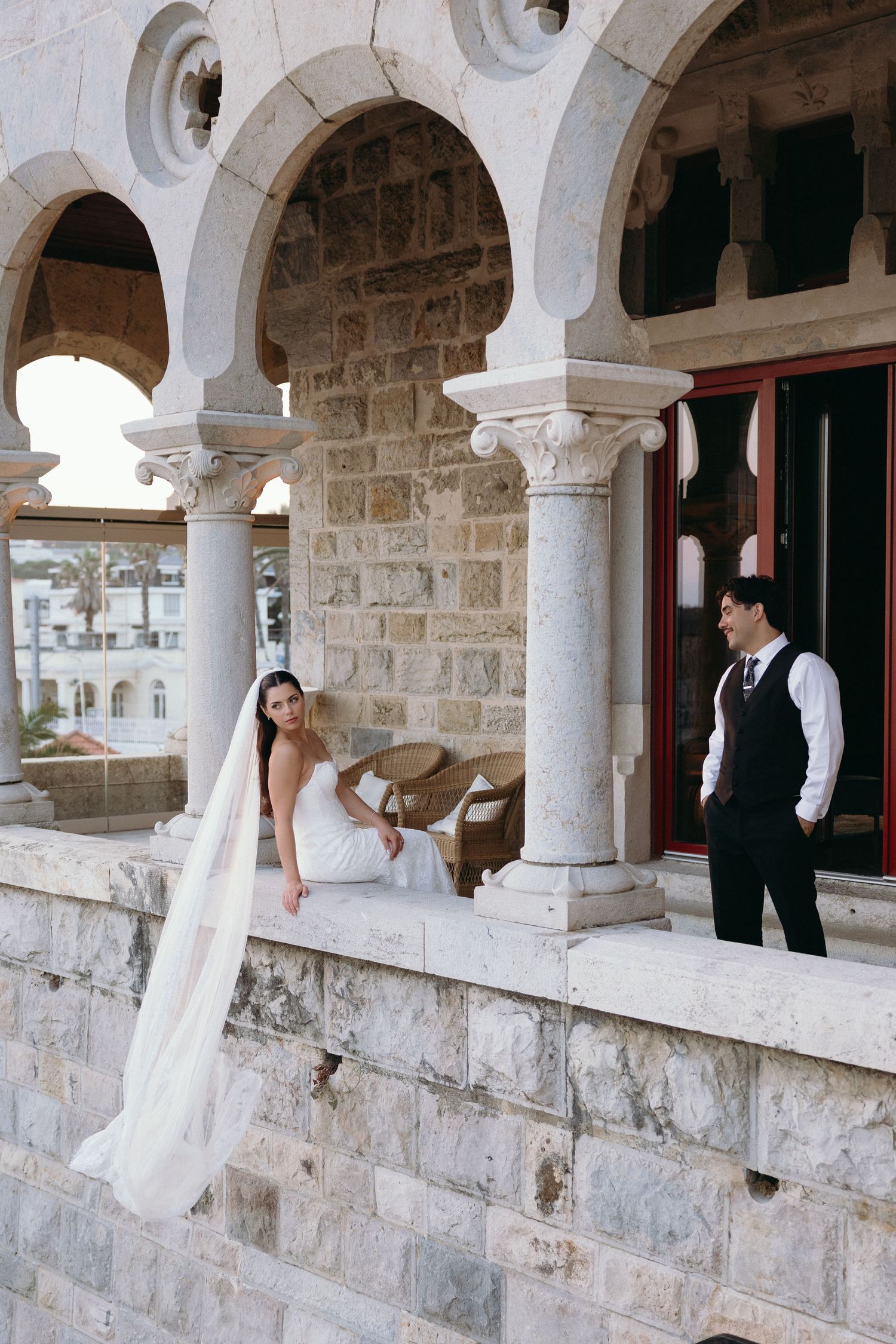 Bride in white gown and veil sits on stone balustrade beside groom in vest at elegant historic stone building.