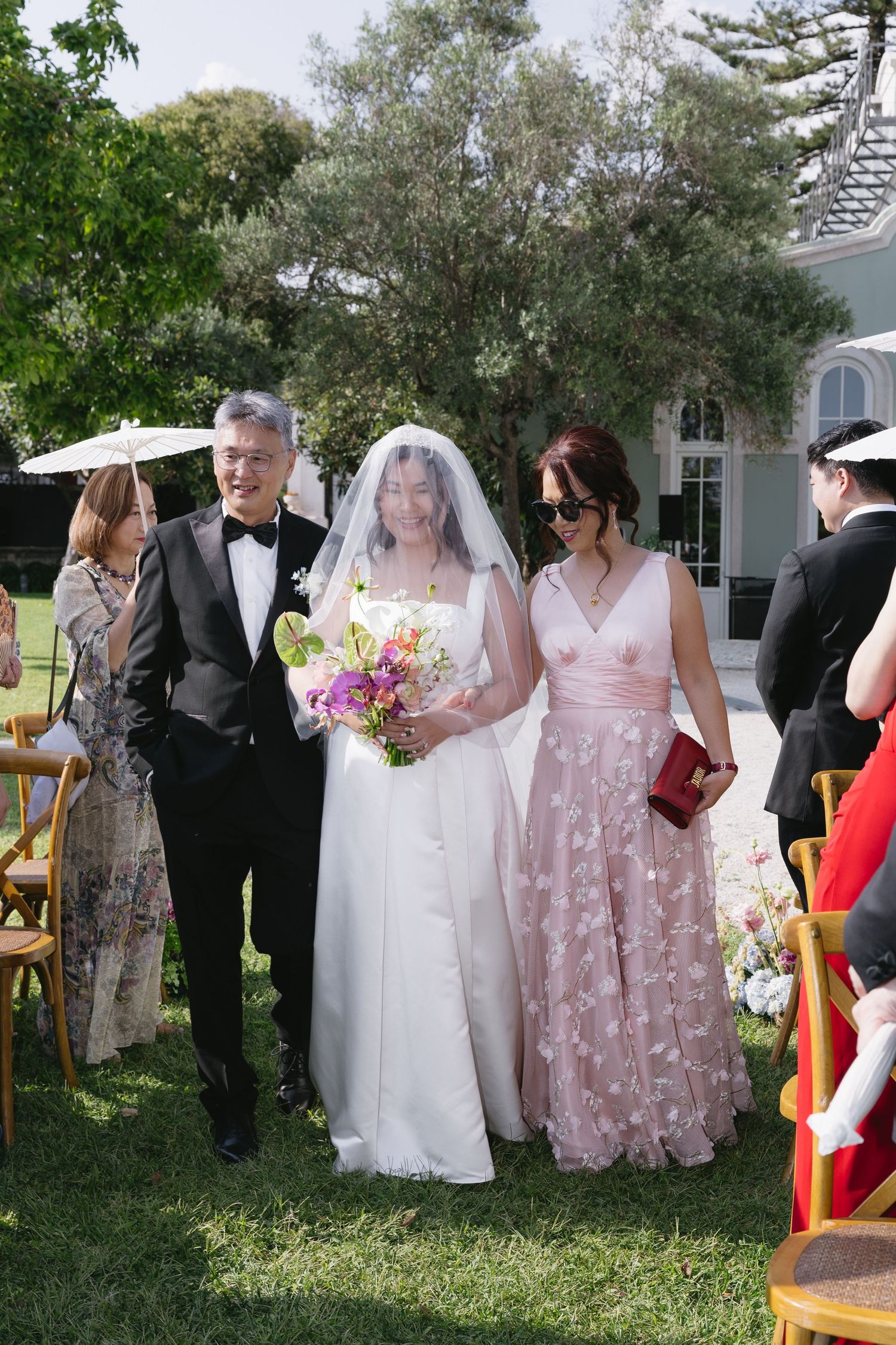 Bride in white gown walks down outdoor aisle with parents in formal attire at a sunny garden wedding ceremony.