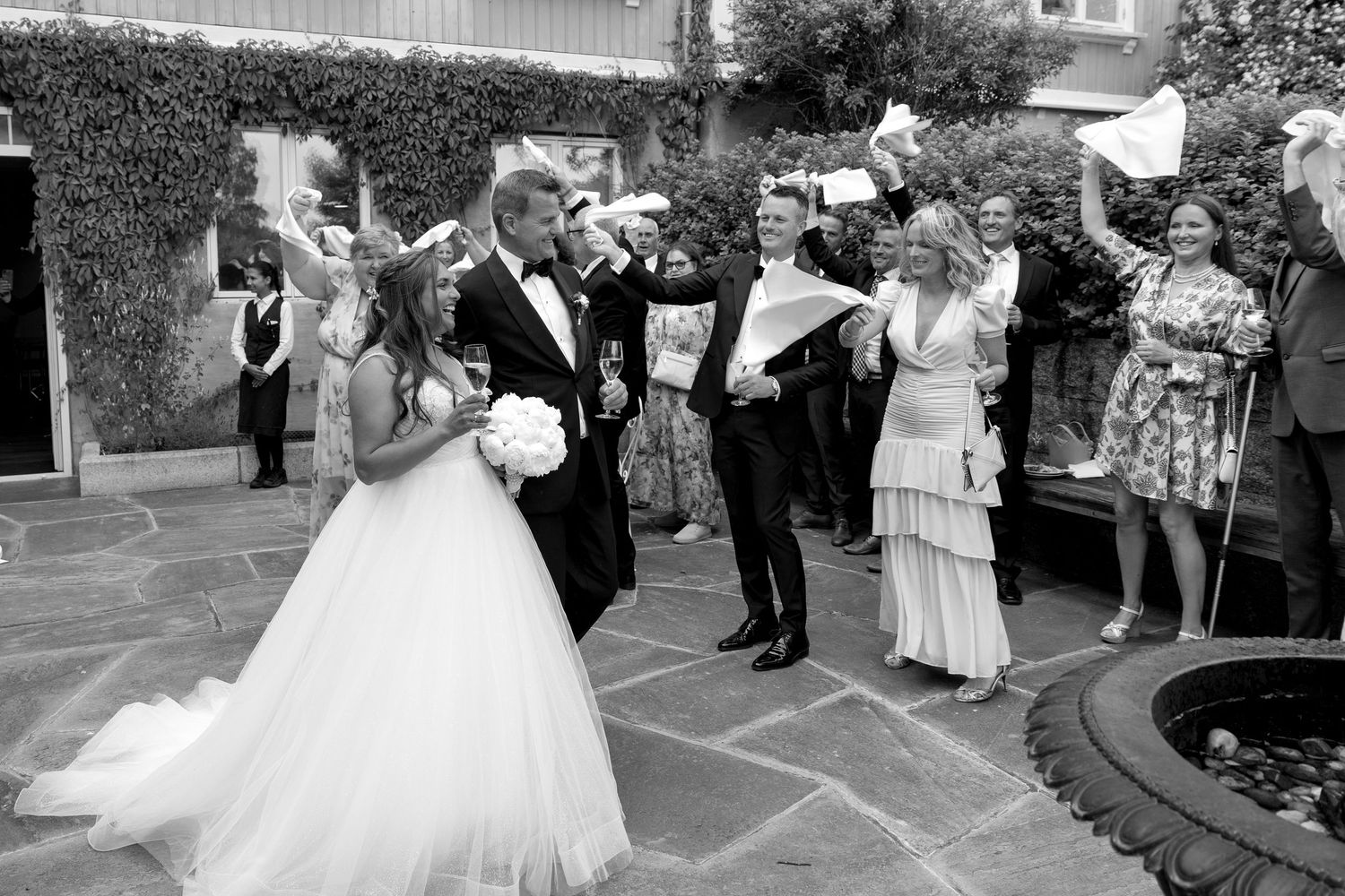 Black and white photo of a bride and groom celebrating with guests tossing paper at an outdoor wedding reception.