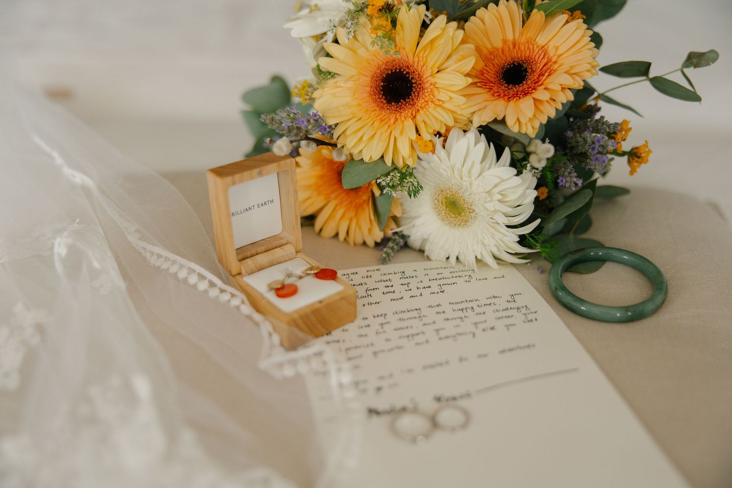 Wedding flat lay with yellow gerbera bouquet, wooden ring box, handwritten vows, and delicate jewelry on lace fabric.