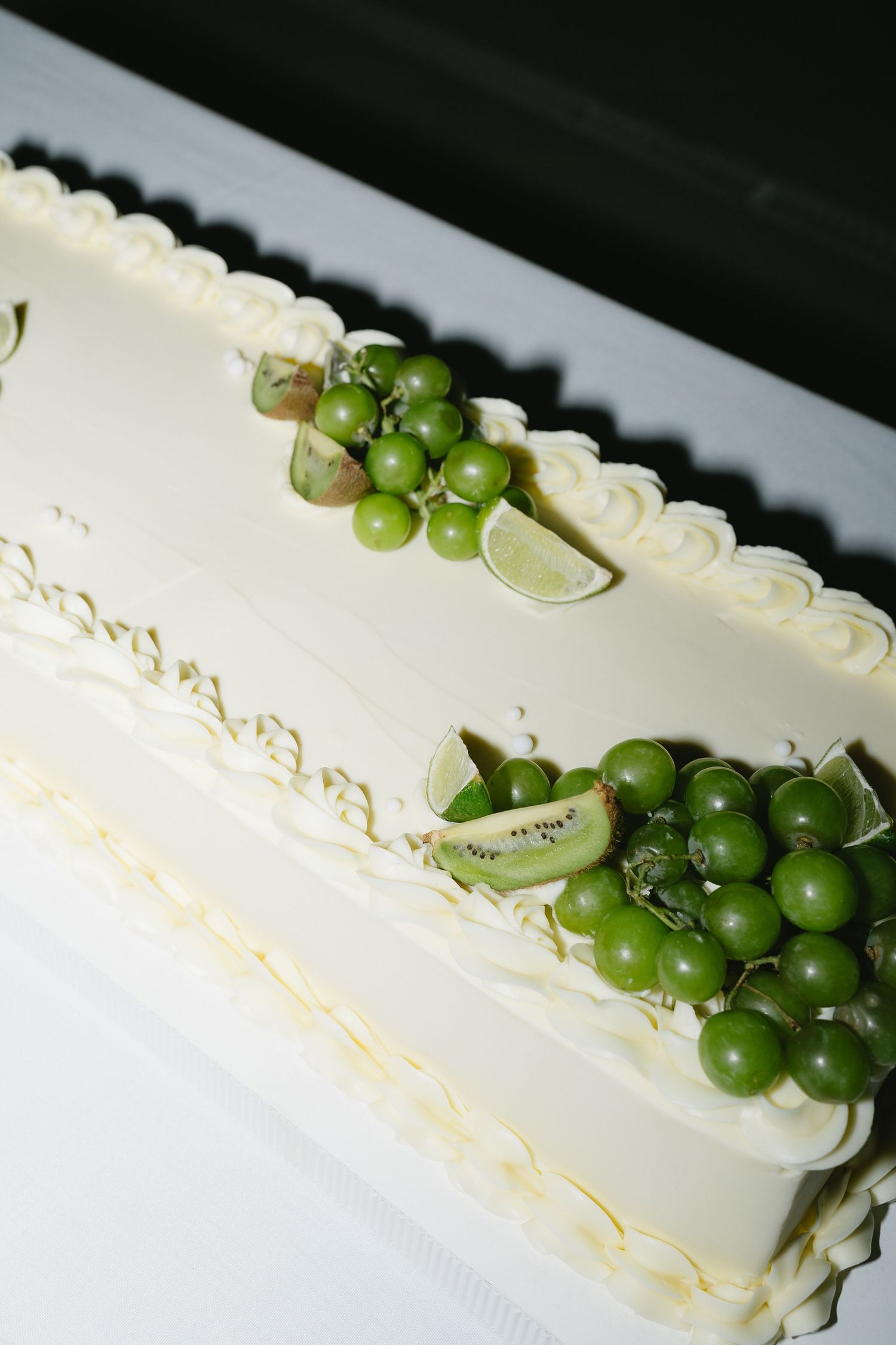 White rectangular wedding cake decorated with green grapes, kiwi slices, and lime garnishes.