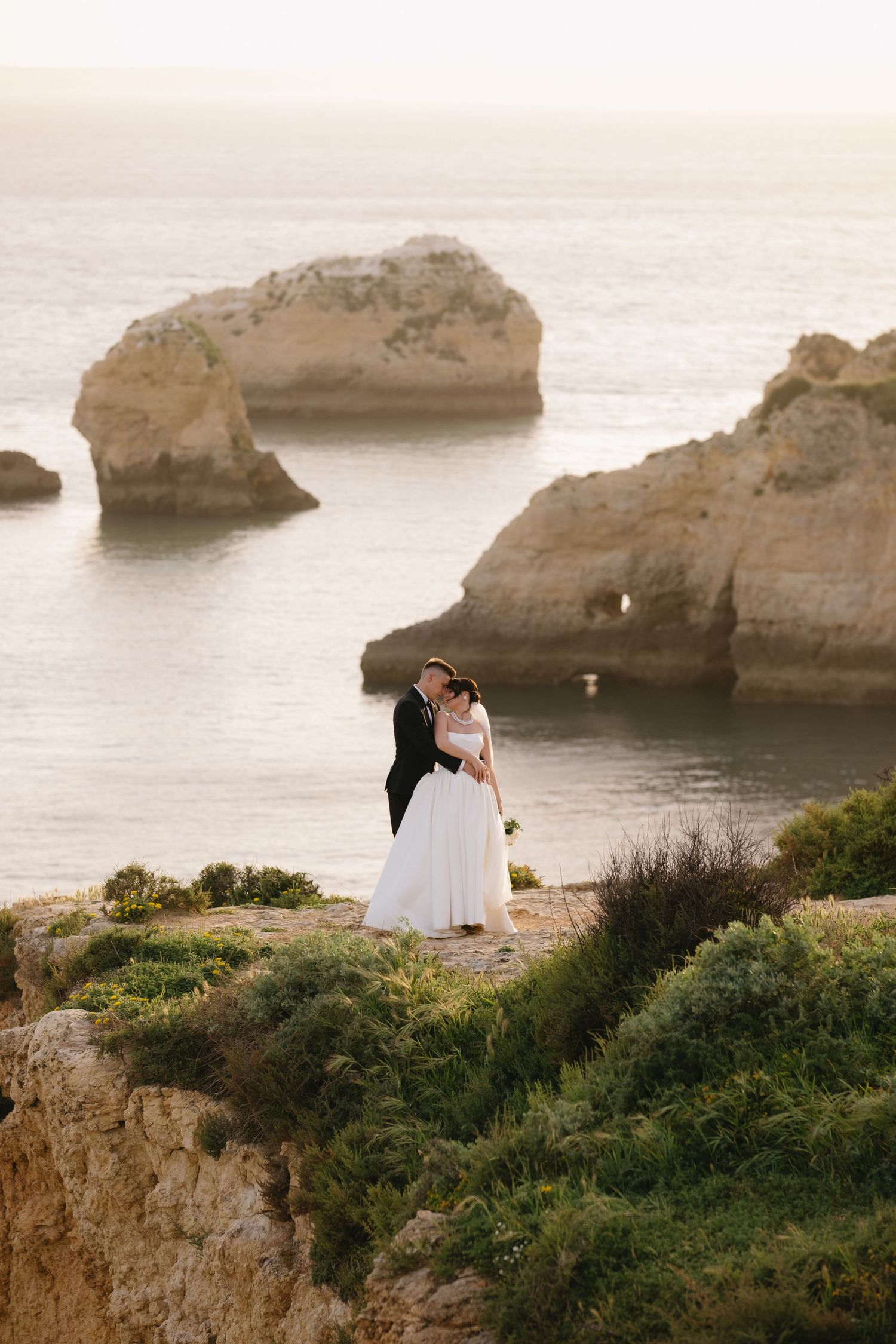 Bride and groom embracing on clifftop overlooking stunning rocky ocean landscape at golden hour.