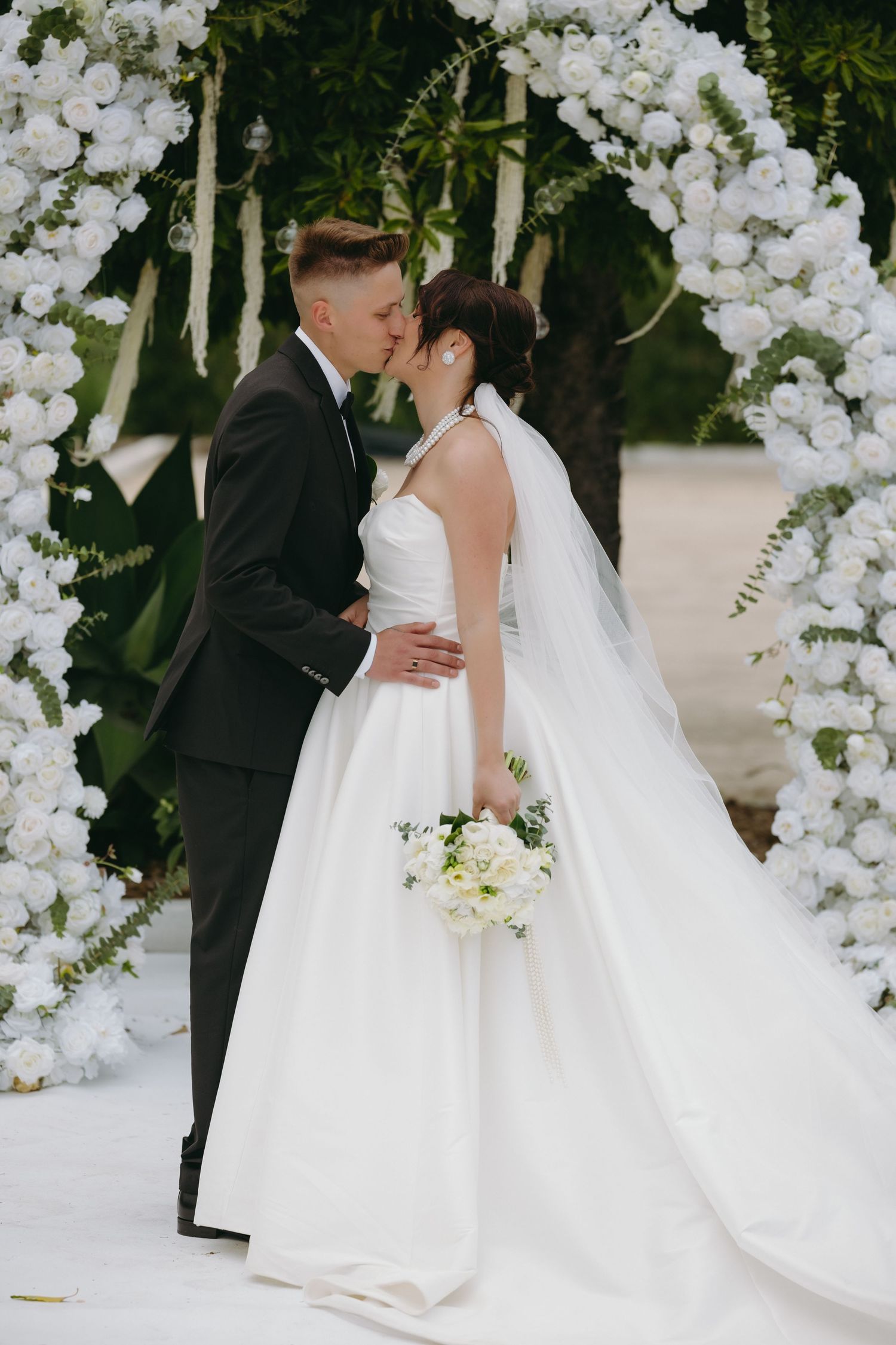 Bride and groom share first kiss under stunning white rose floral arch at outdoor wedding ceremony.