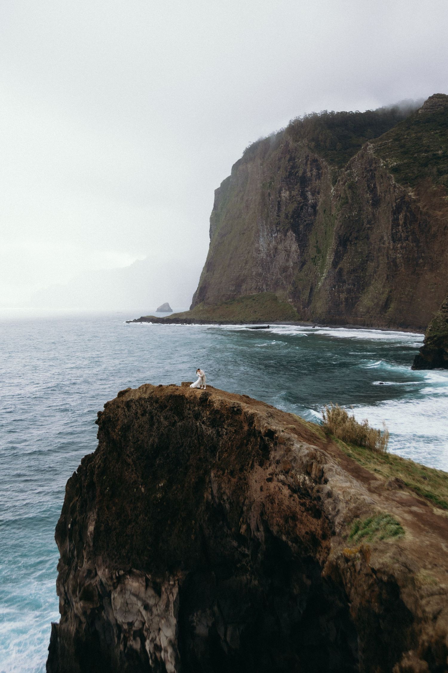 Lone wedding couple sitting on a dramatic rocky cliff edge overlooking the vast ocean and towering cliffs in Madeira.