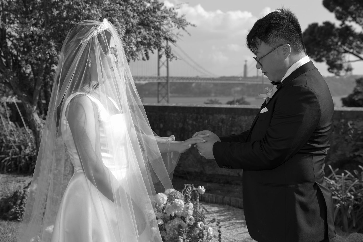 Groom placing wedding ring on bride's finger during outdoor ceremony, black and white photo with bridge in background.