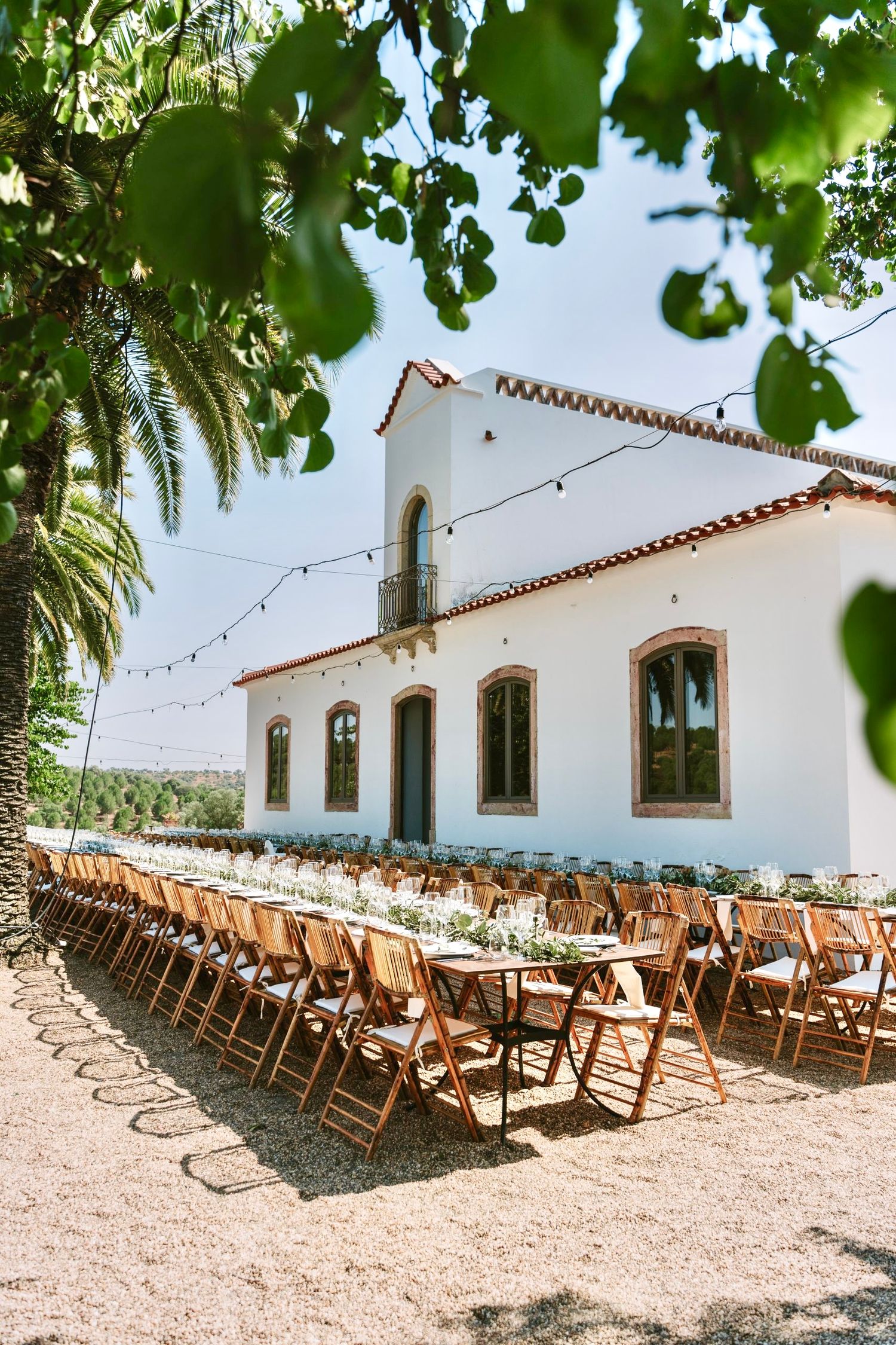 Elegant outdoor wedding banquet with long wooden tables beside a white Portuguese quinta under string lights.