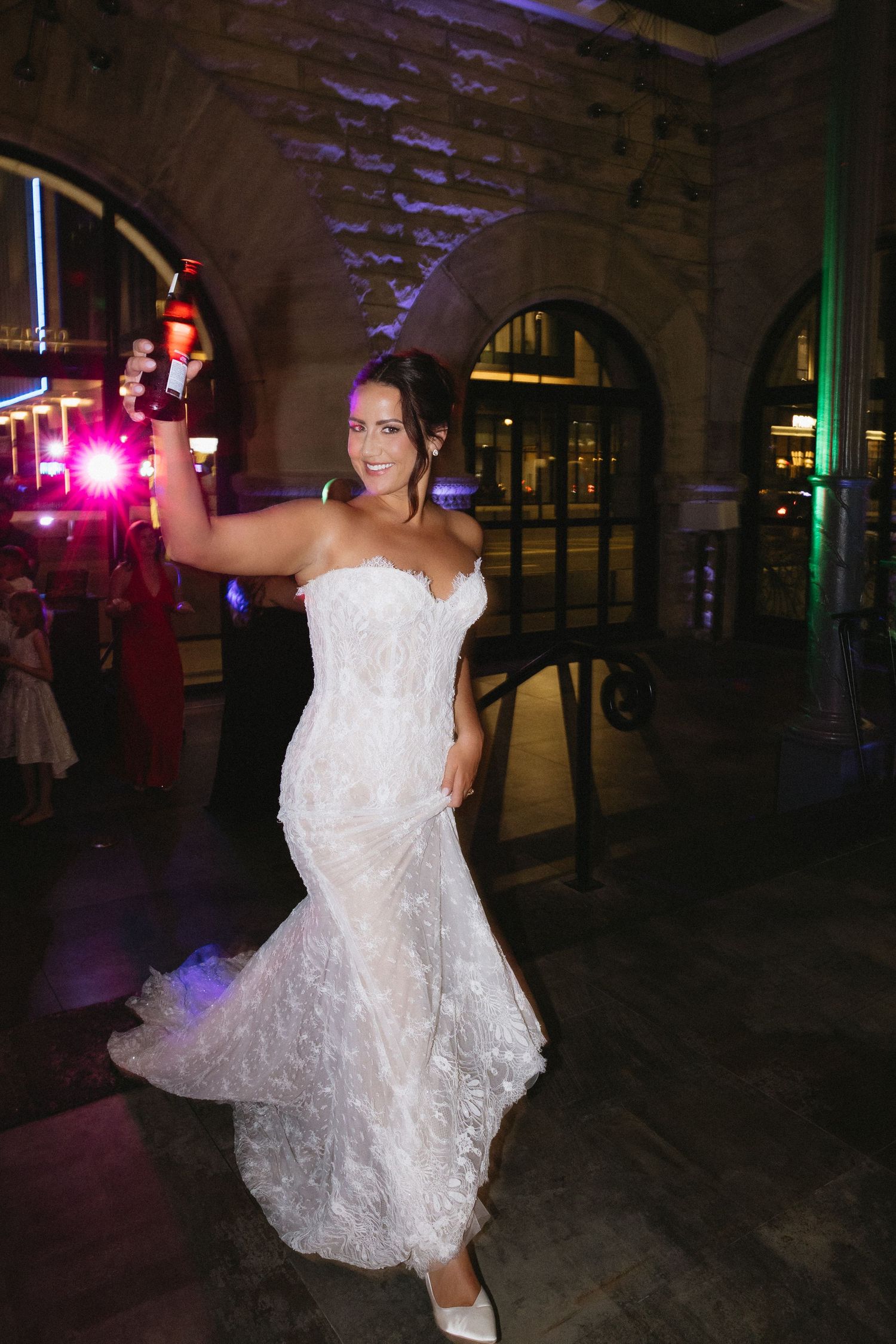 Bride in lace mermaid gown dancing and holding champagne bottle at elegant evening wedding reception.